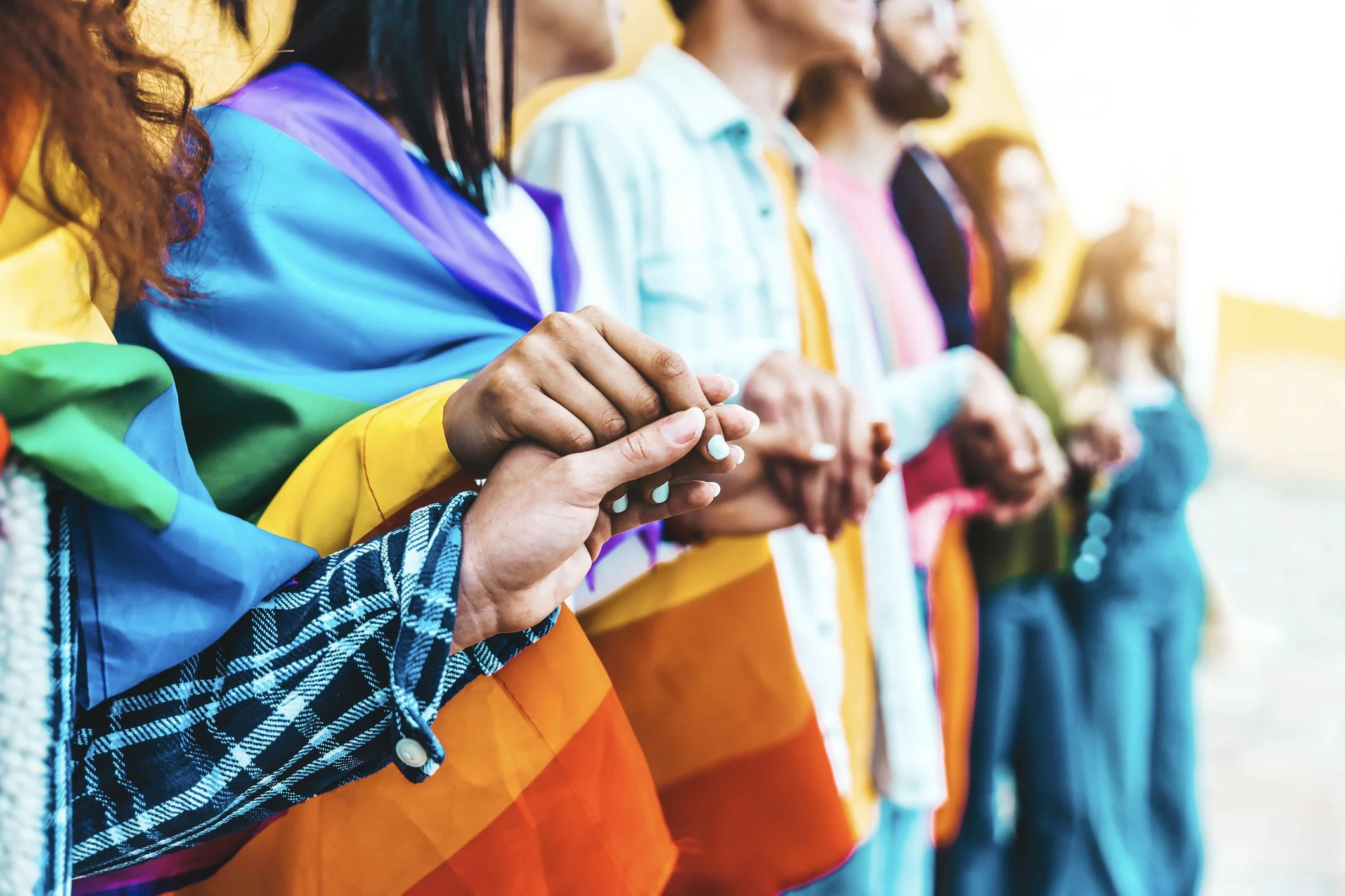 Group of diverse people holding hands, wearing rainbow-colored clothing, standing in a line outdoors during sunset.