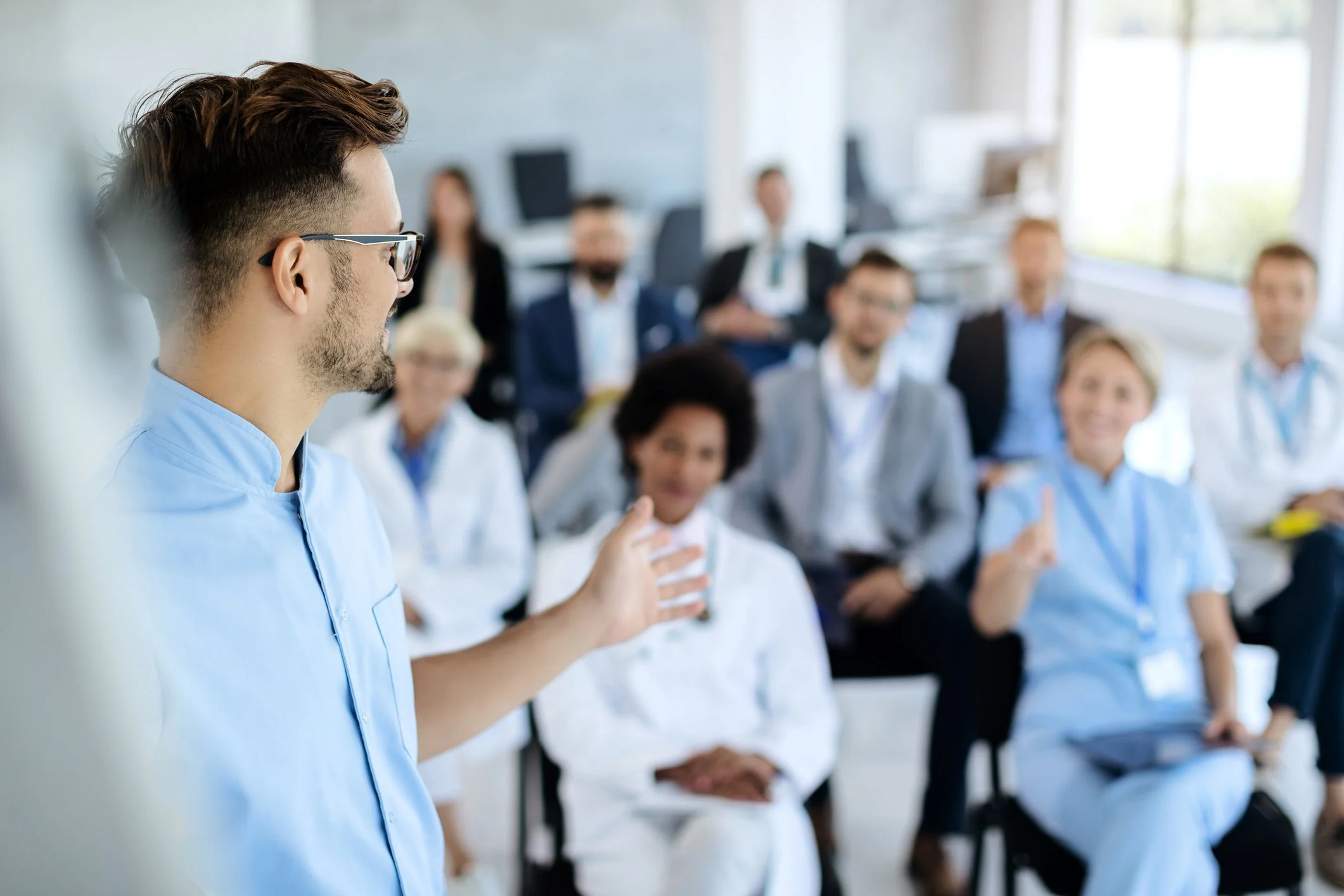 A man giving a presentation to a group of healthcare professionals, including doctors and nurses, in a brightly lit conference room.