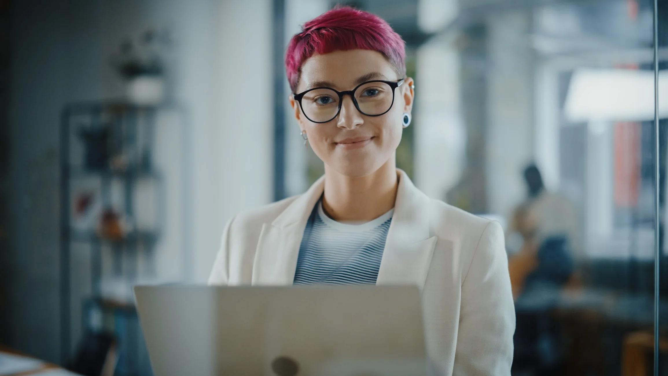 A young woman with pink hair, glasses, and earrings working on a laptop in a bright office space.