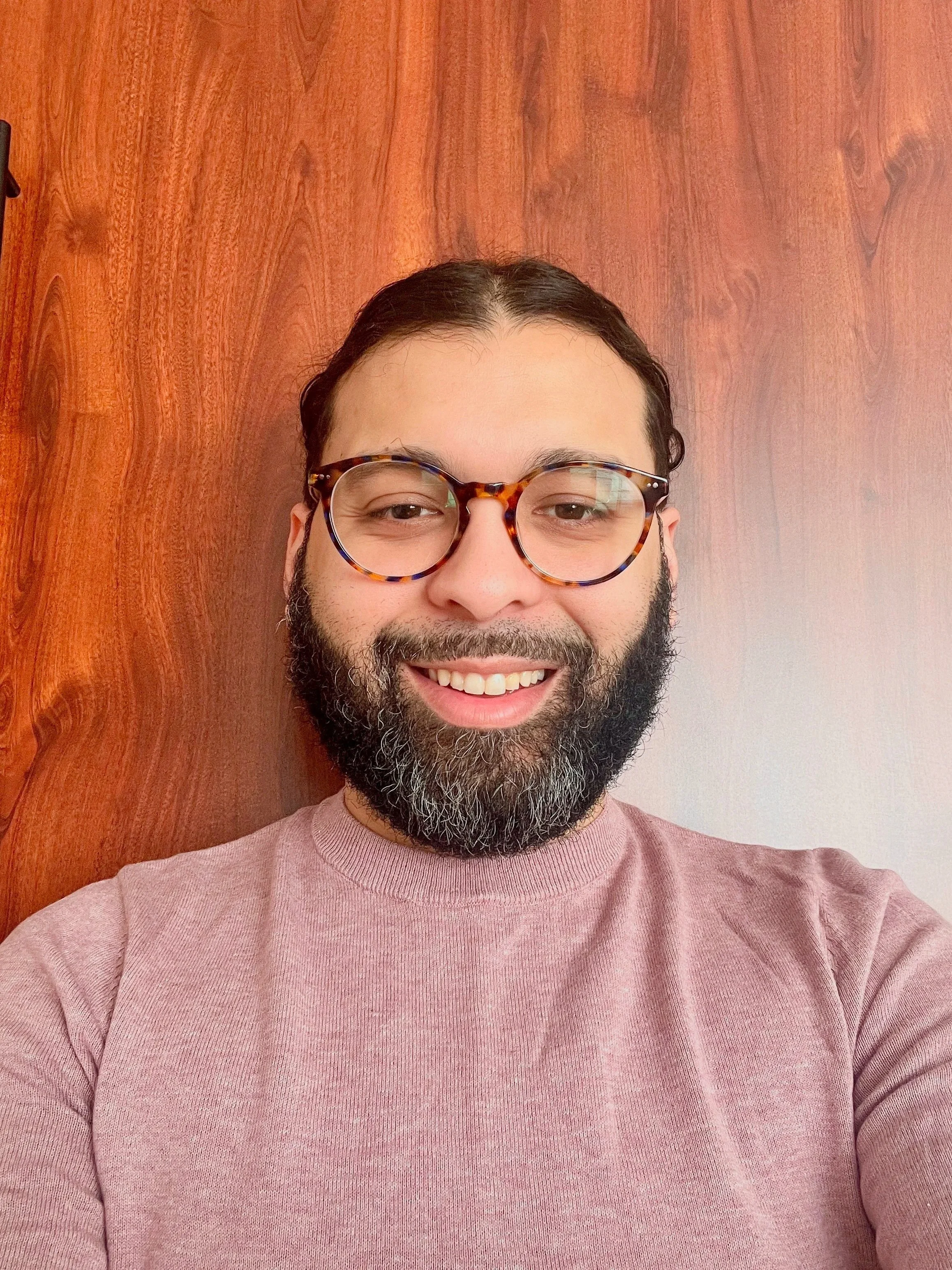 A man with glasses and a beard smiling, standing in front of a wooden wall.