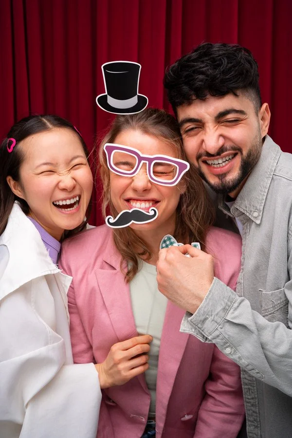 Three people smiling and having fun at a photo booth with props including a top hat, glasses, and a moustache, against a red curtain background.