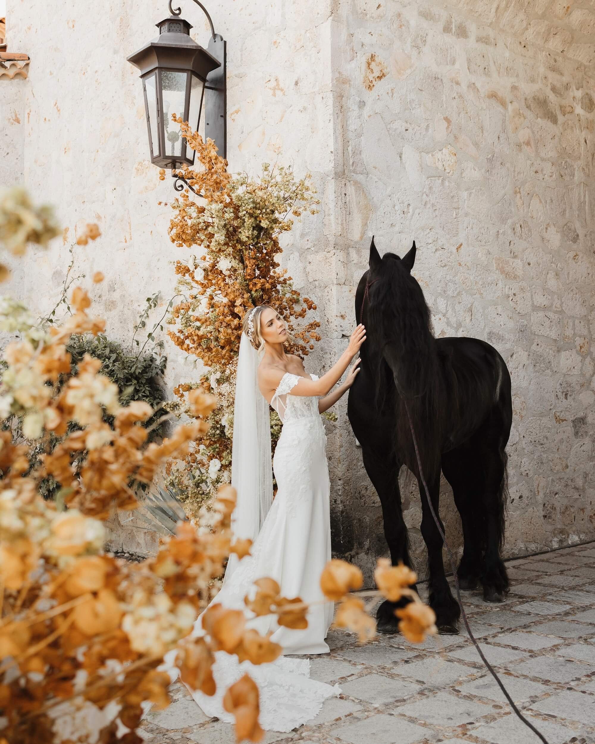 Bride in a white wedding dress touching a black horse by an old stone wall with orange flowers and a lantern.
