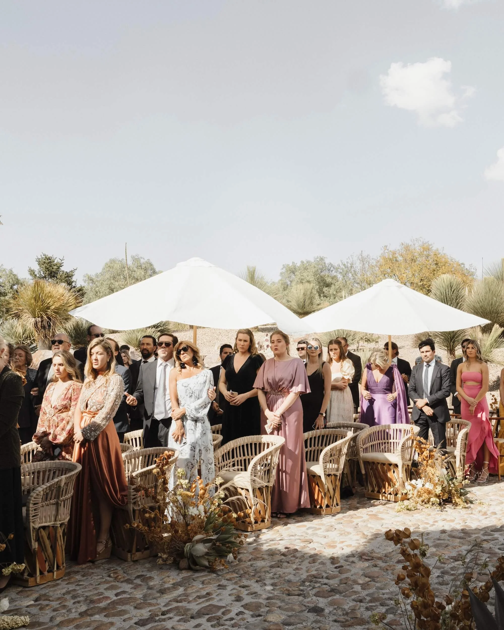 Group of people attending an outdoor wedding ceremony, standing on cobblestone paving with white umbrellas and desert plants in the background