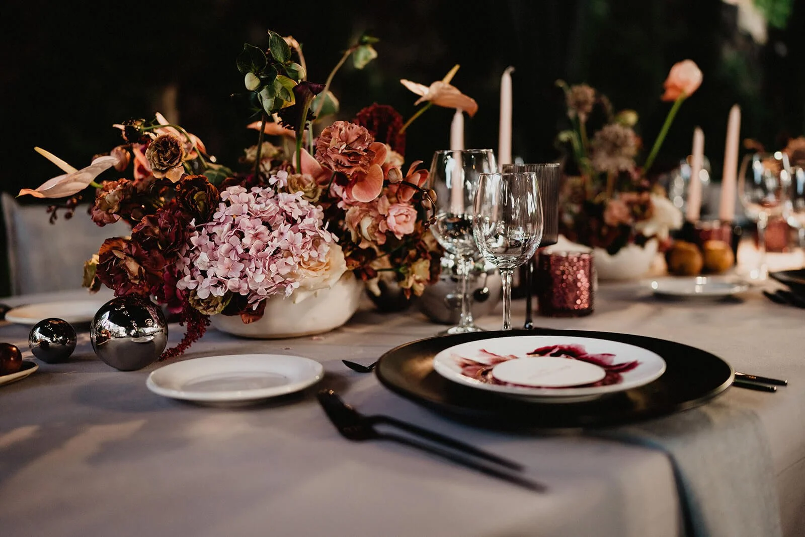 Elegant dining table setup with pink and purple floral centerpiece, wine glasses, white and black plates, candles, and decorative ornaments.