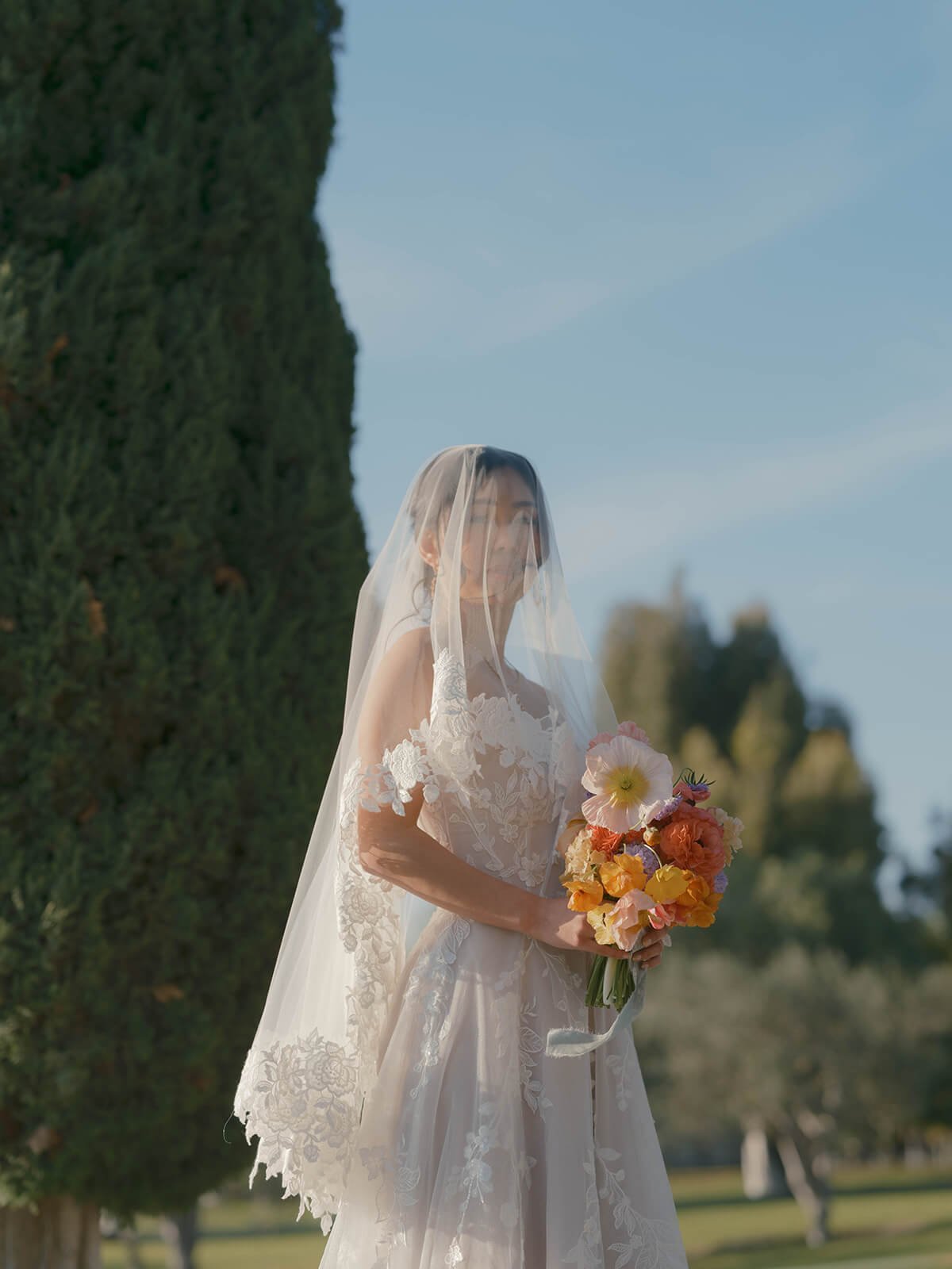 A woman in a white lace wedding dress holding a colorful bouquet of flowers, standing outdoors with a large green tree and blue sky in the background, wearing a veil over her face.