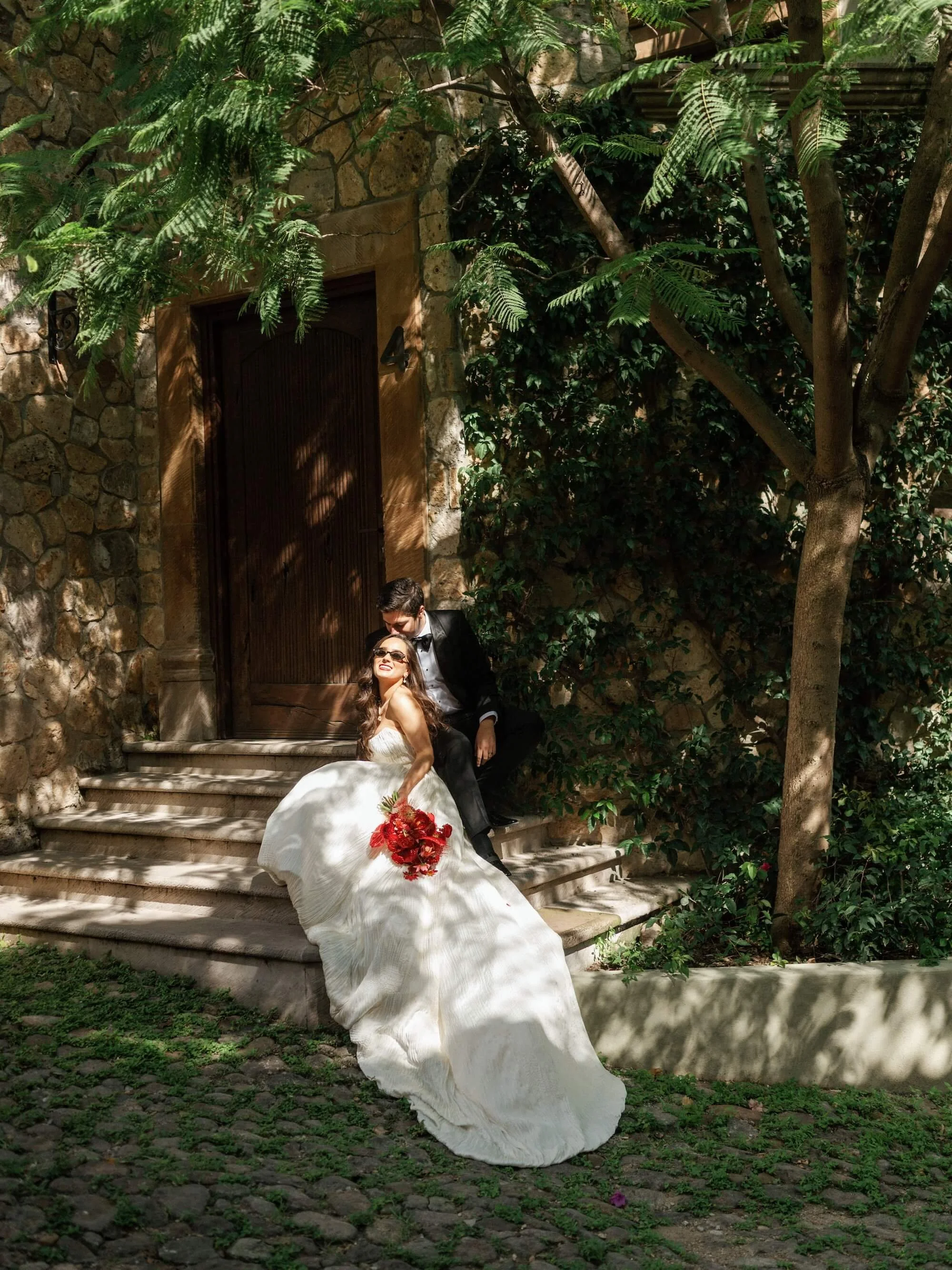 A bride in a white wedding gown holding a bouquet of red flowers, sitting on steps at a stone building, with a groom in a black tuxedo sitting behind her. They are outdoors in sunny, shaded surroundings with greenery.