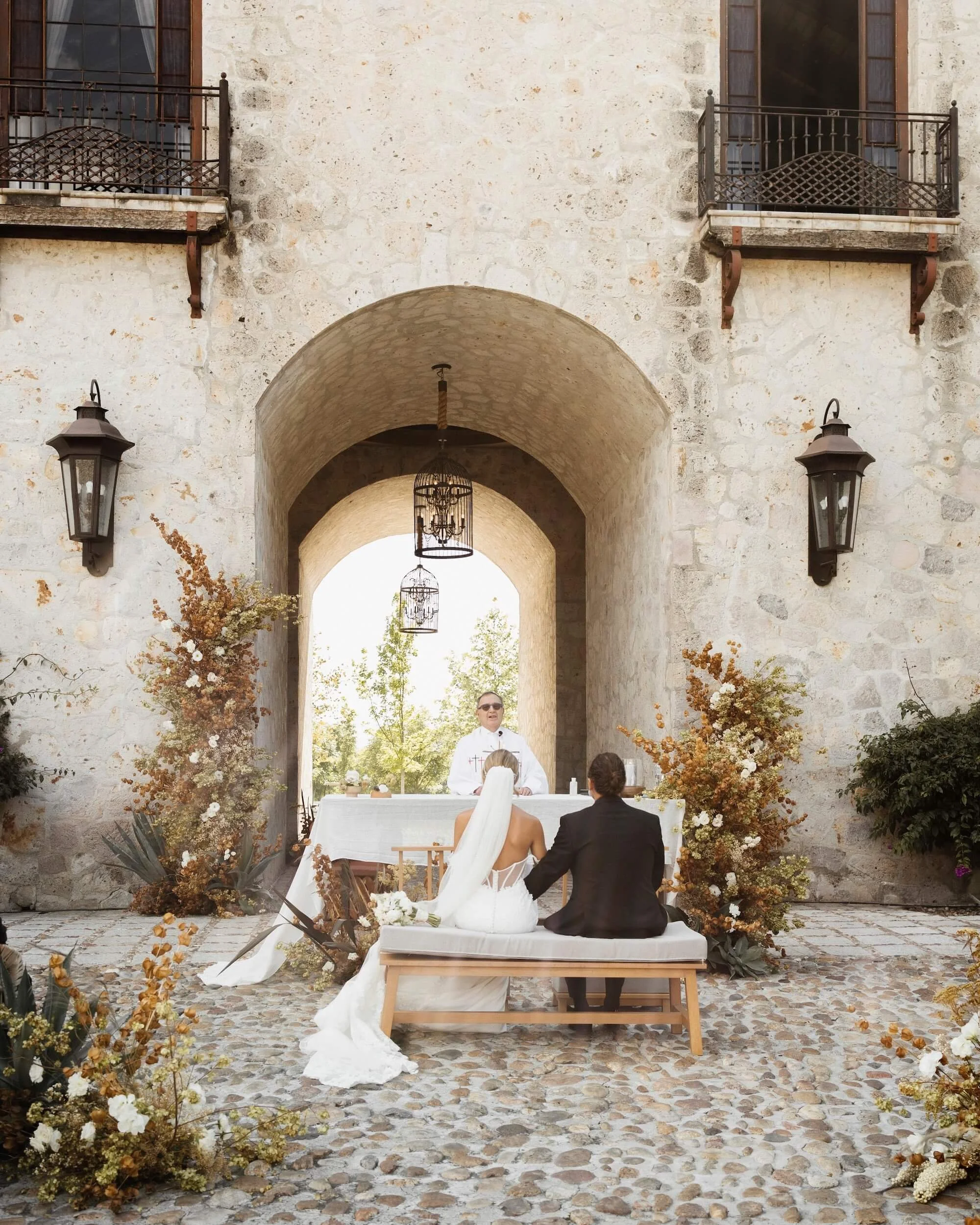 A couple in wedding attire sitting on a bench at an outdoor wedding ceremony in front of an officiant, with decorative plants and an arched stone wall background.