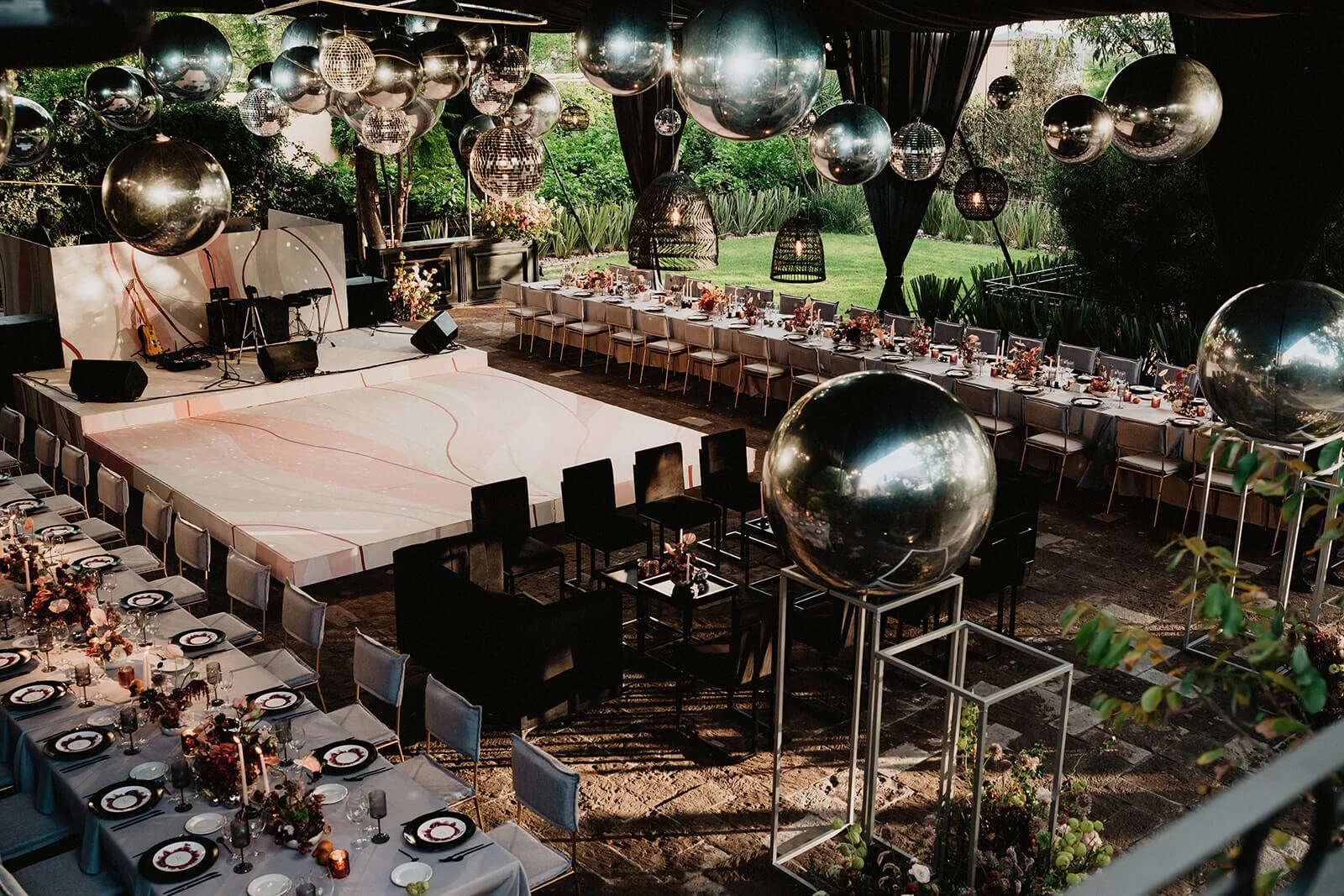 Decorated outdoor event space with long dining tables, floral centerpieces, large reflective balloons, a stage with musical instruments, and a lush green garden in the background.