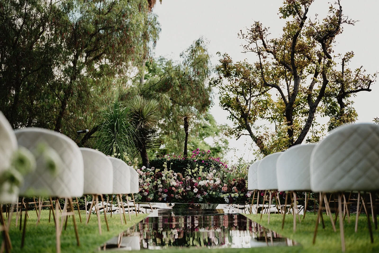 Outdoor event setup with white cushioned chairs arranged along a grassy aisle, floral arch with pink and white flowers, and tall trees in the background.