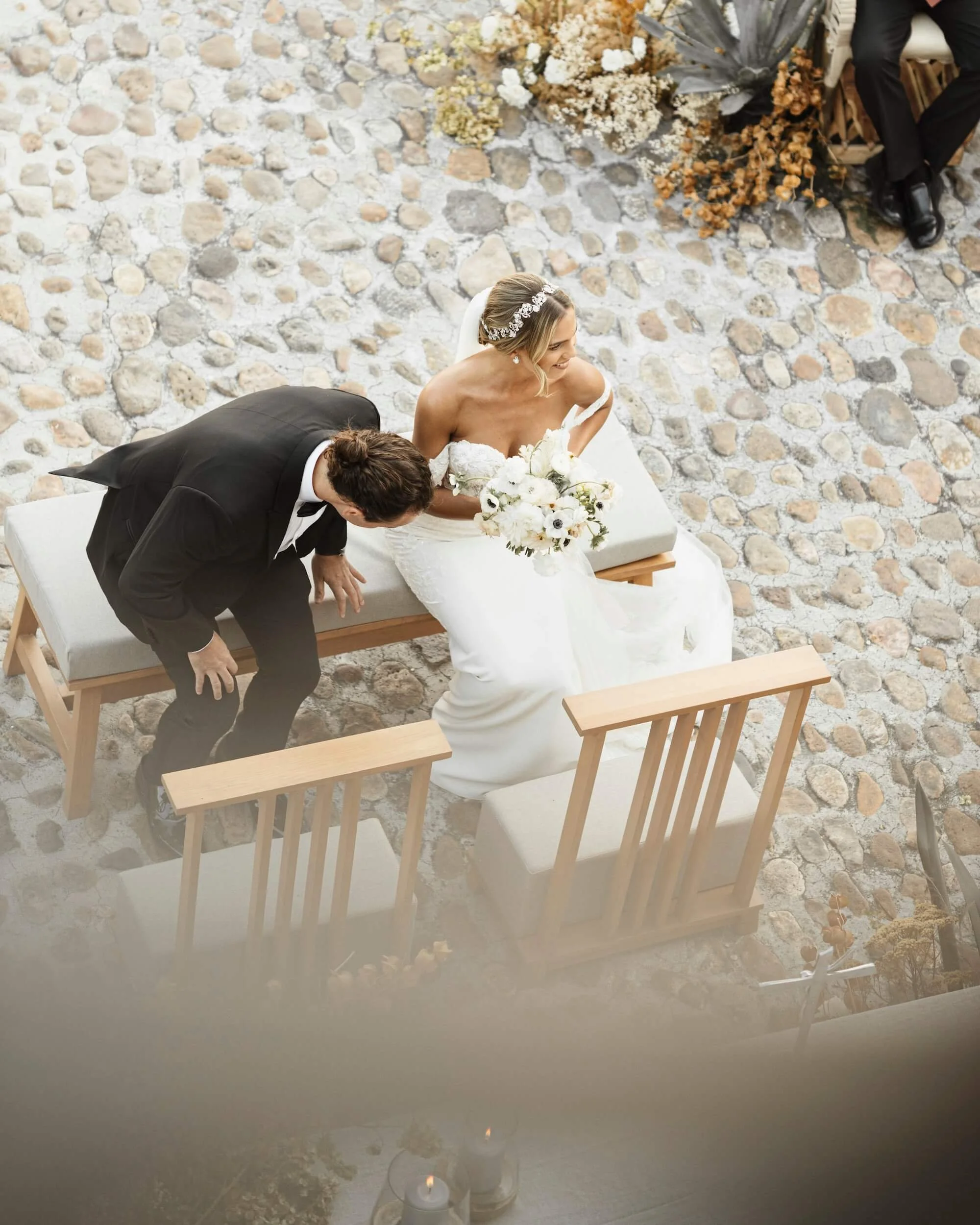A bride in a white wedding dress holding a bouquet of white flowers, seated on a bench. A man in a black suit leans over to speak to her. They are in a venue with stone flooring and wooden chairs, decorated with flowers and candles.