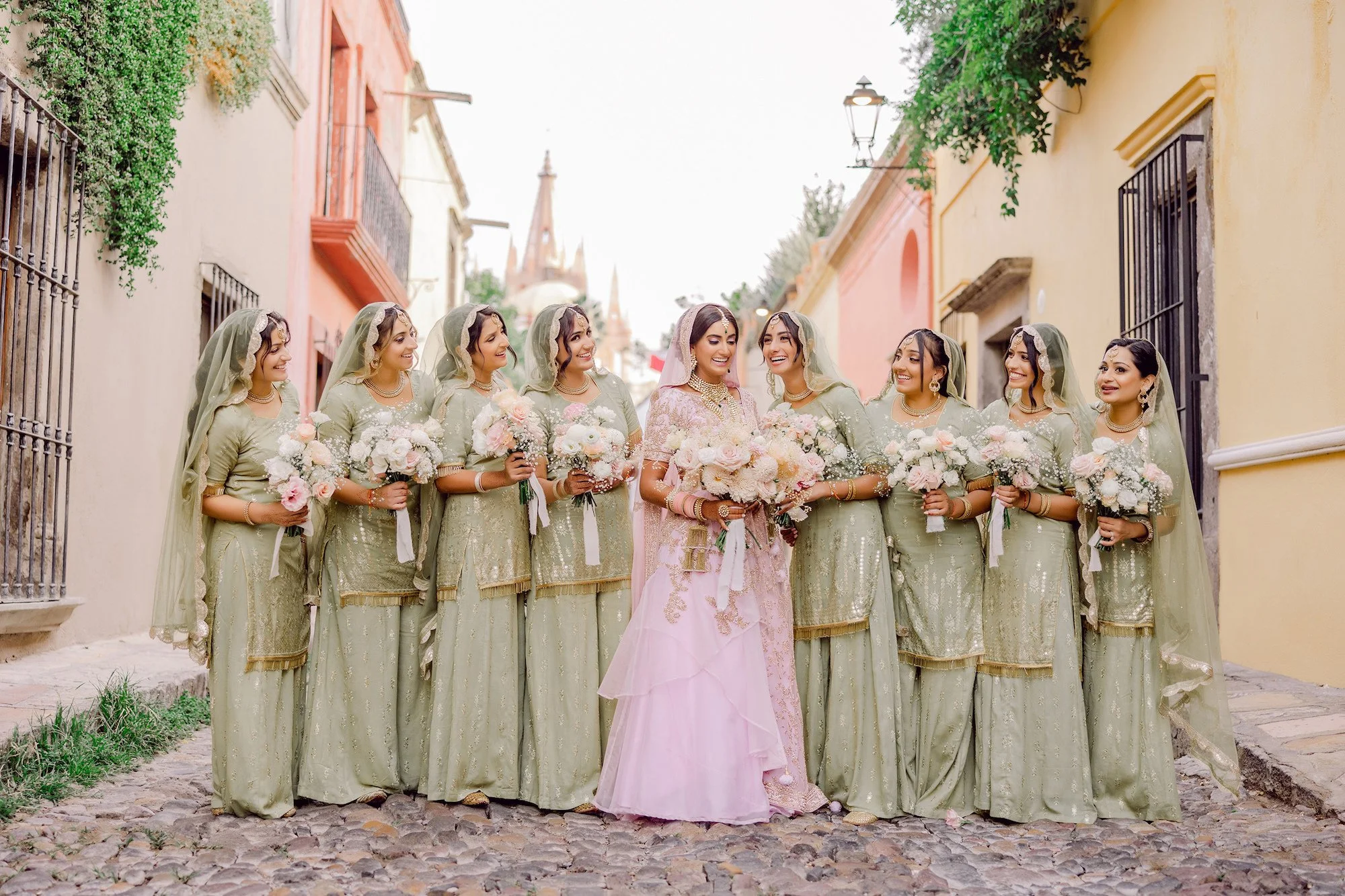 A bride in pink traditional wedding dress with bridesmaids in matching light green dresses, standing in a colorful street, holding bouquets, smiling, during a wedding celebration.