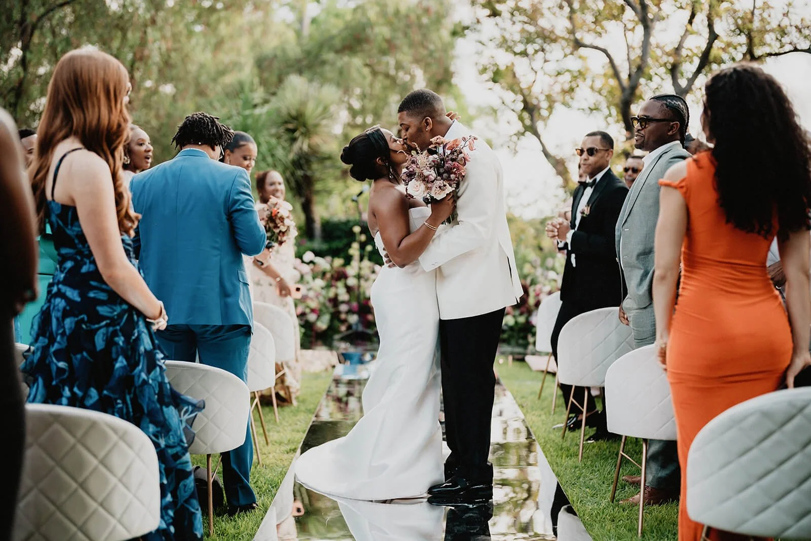 A newly married couple sharing a kiss during an outdoor wedding ceremony, with guests standing on either side of the aisle, trees, and floral decorations in the background.