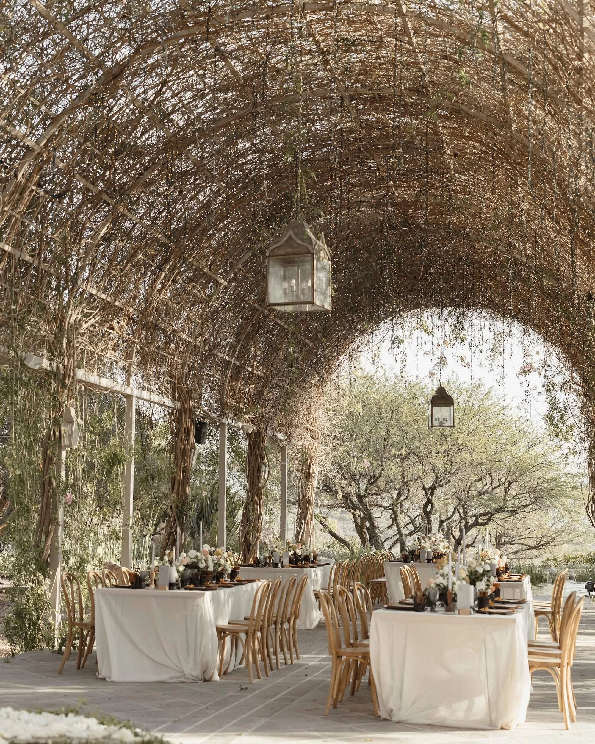 Outdoor wedding reception under a rustic wooden arbor with hanging lanterns, long tables with white tablecloths, floral centerpieces, and wooden chairs, surrounded by trees and natural scenery.