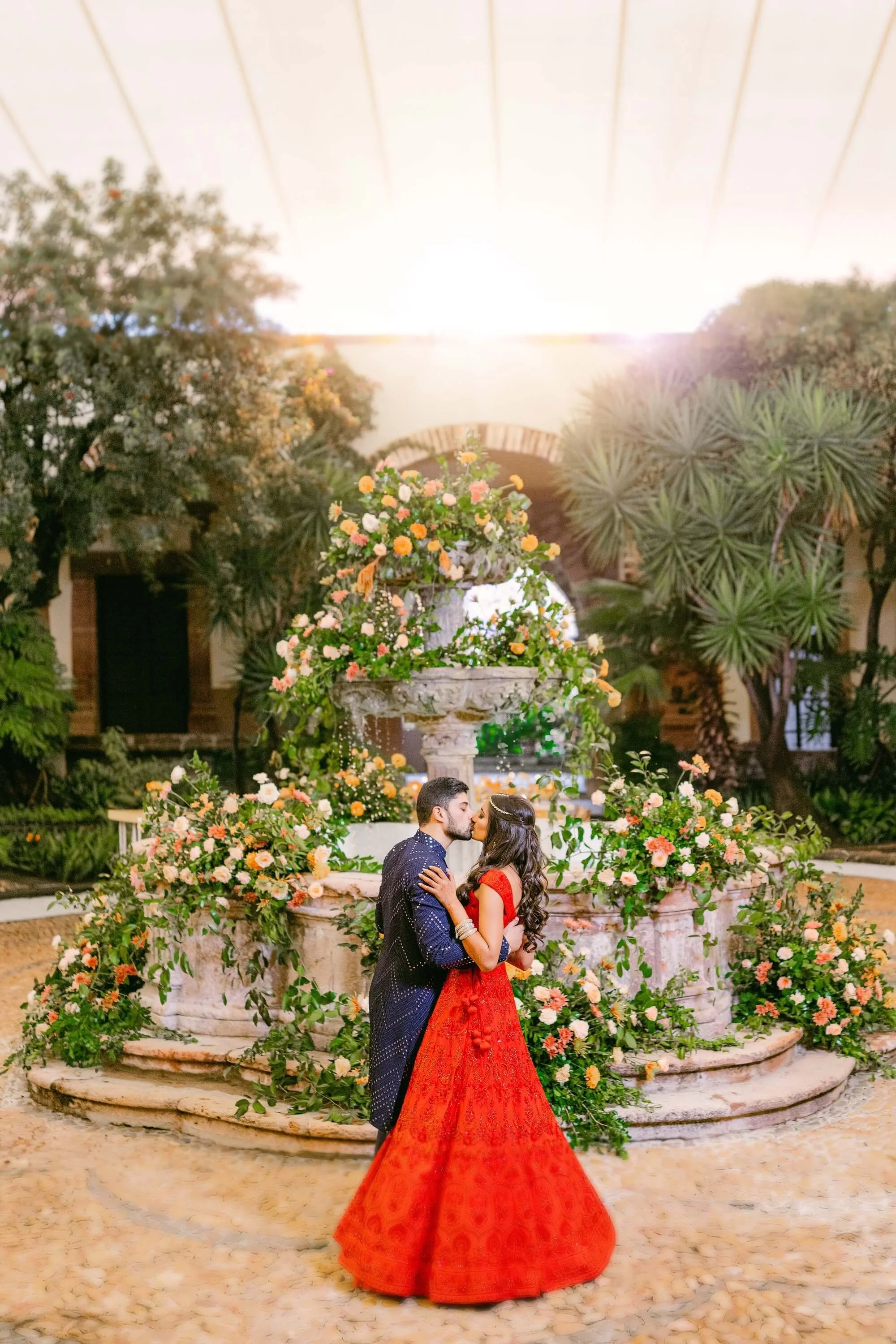 A couple is kissing in front of a floral fountain during sunset in an outdoor garden.