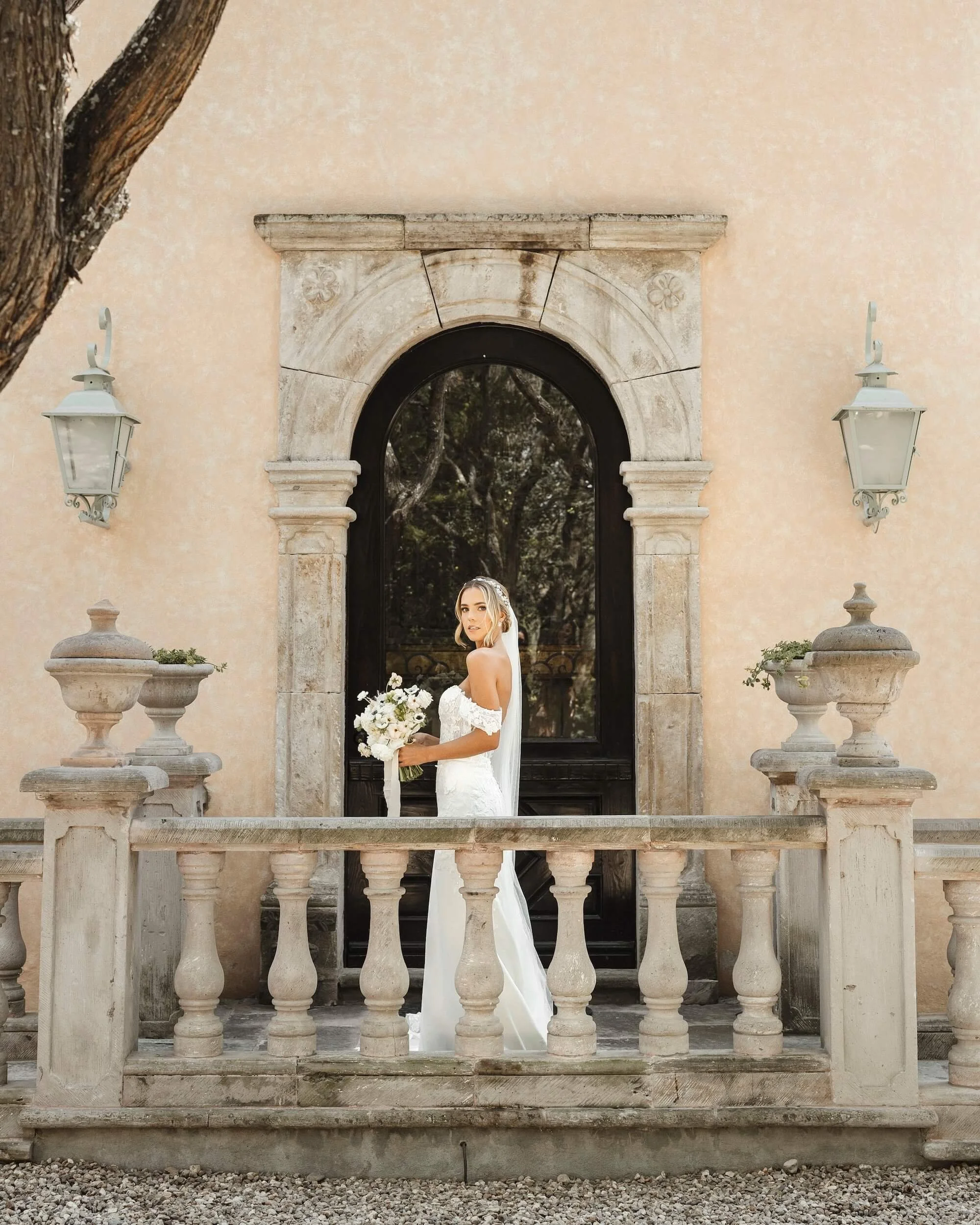 A bride in a white wedding dress standing on a stone balcony holding a bouquet, with a large arched window and beige wall behind her, and trees visible through the window.