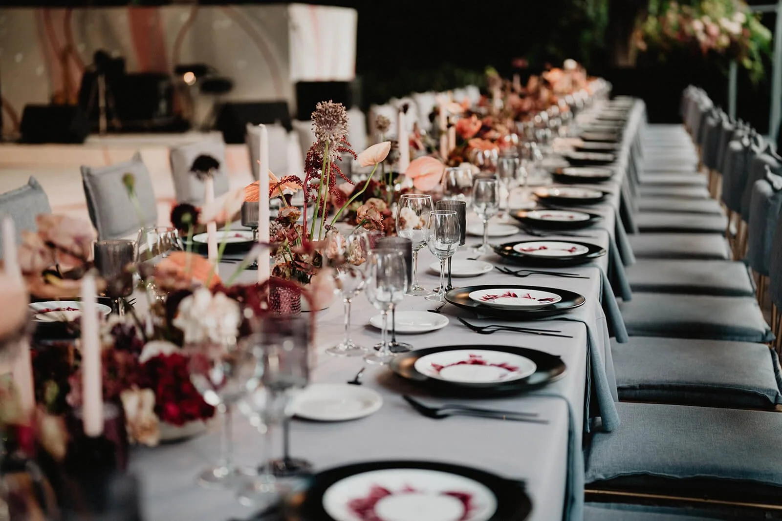 Long banquet table set for a formal event with plates, glasses, black napkins, and pink flowers with candles as centerpieces.