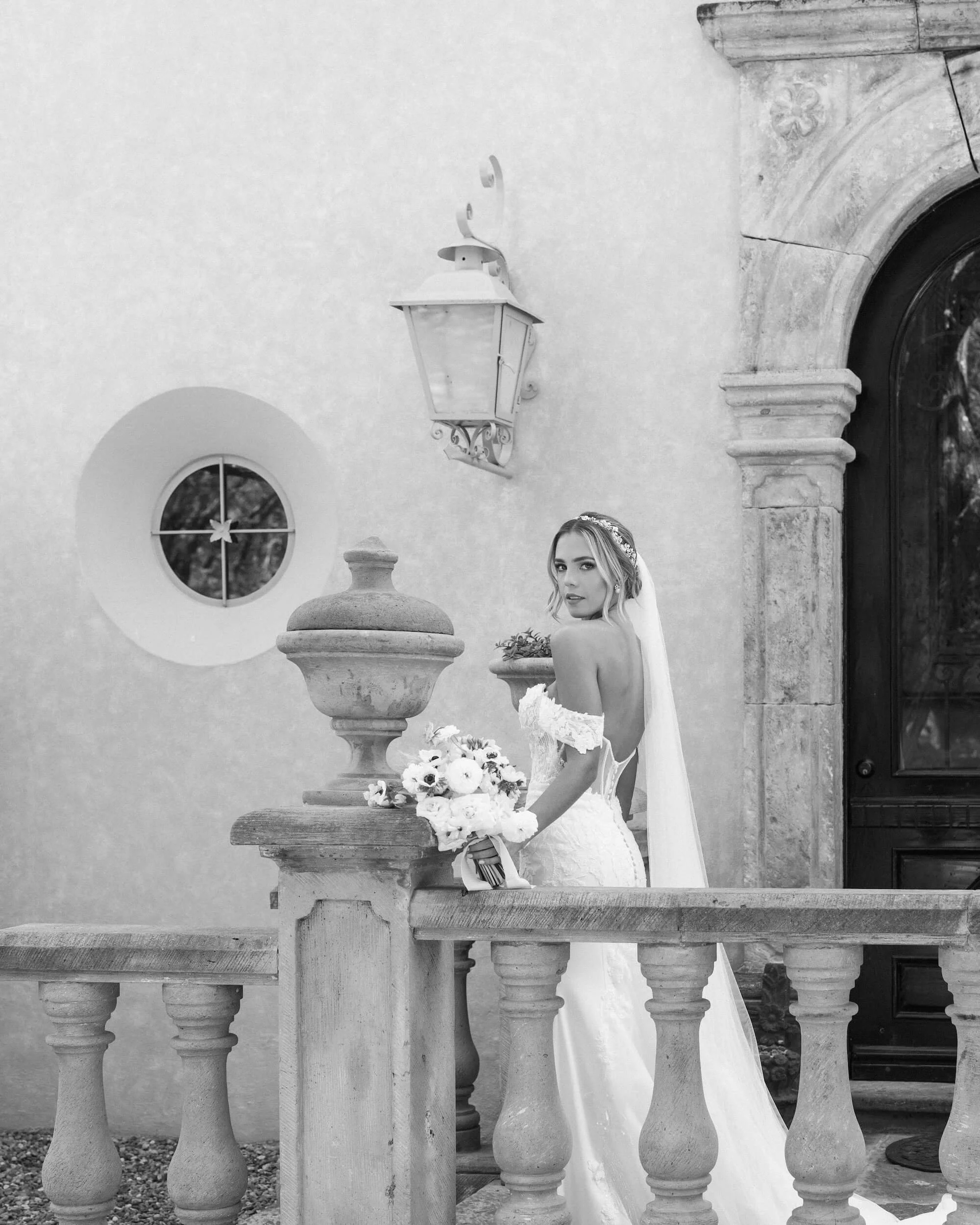 Black and white photo of a bride in a wedding dress standing on a stone balcony, holding a bouquet, with a stone wall, an arched window, and a lantern above her