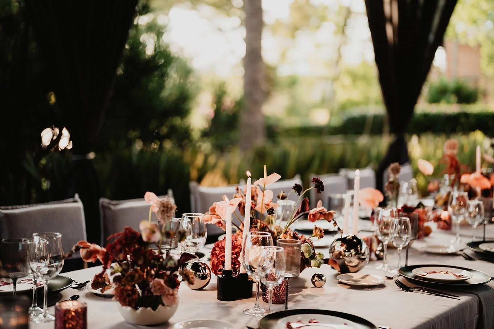 Formal outdoor dinner table decorated with pink and red flowers, candles, and glassware under a tent with trees in the background.