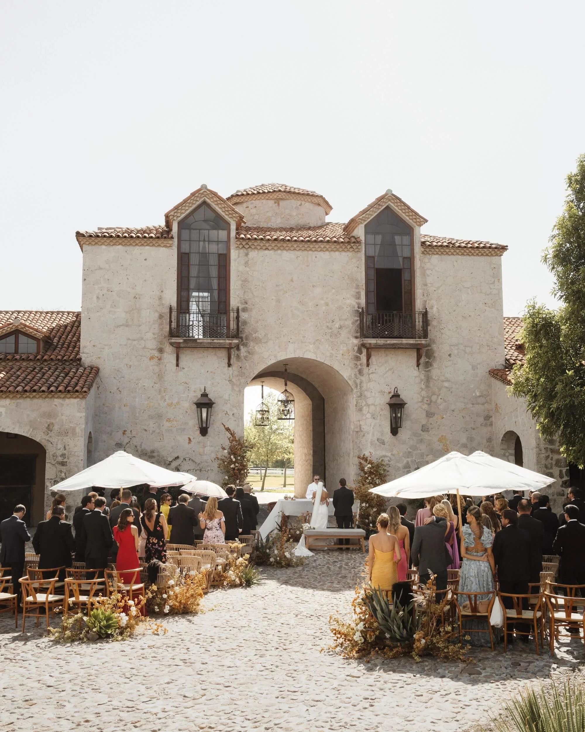 Wedding ceremony outside a stone building with arched entrance, guests seated under umbrellas, bride and groom at the altar.