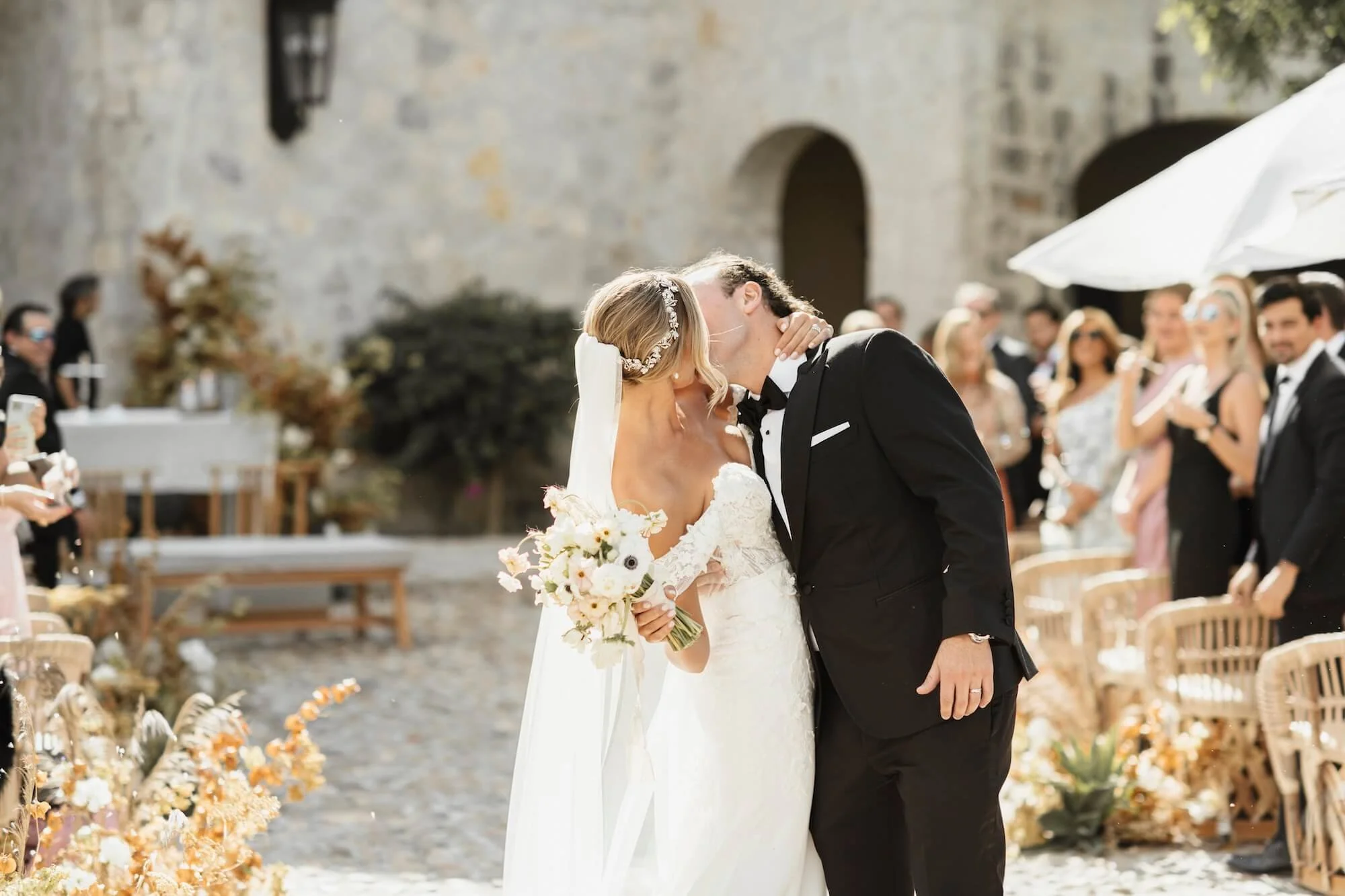 A bride and groom share a kiss during their outdoor wedding, with guests watching in the background.