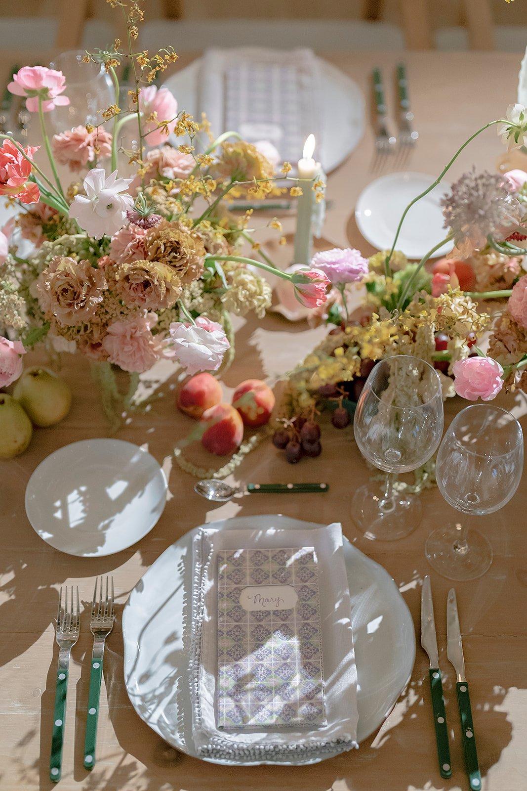 A formal dining table decorated with pink, white, and beige flowers, peach fruits, and wine glasses, set with plates, cutlery, and a menu card.