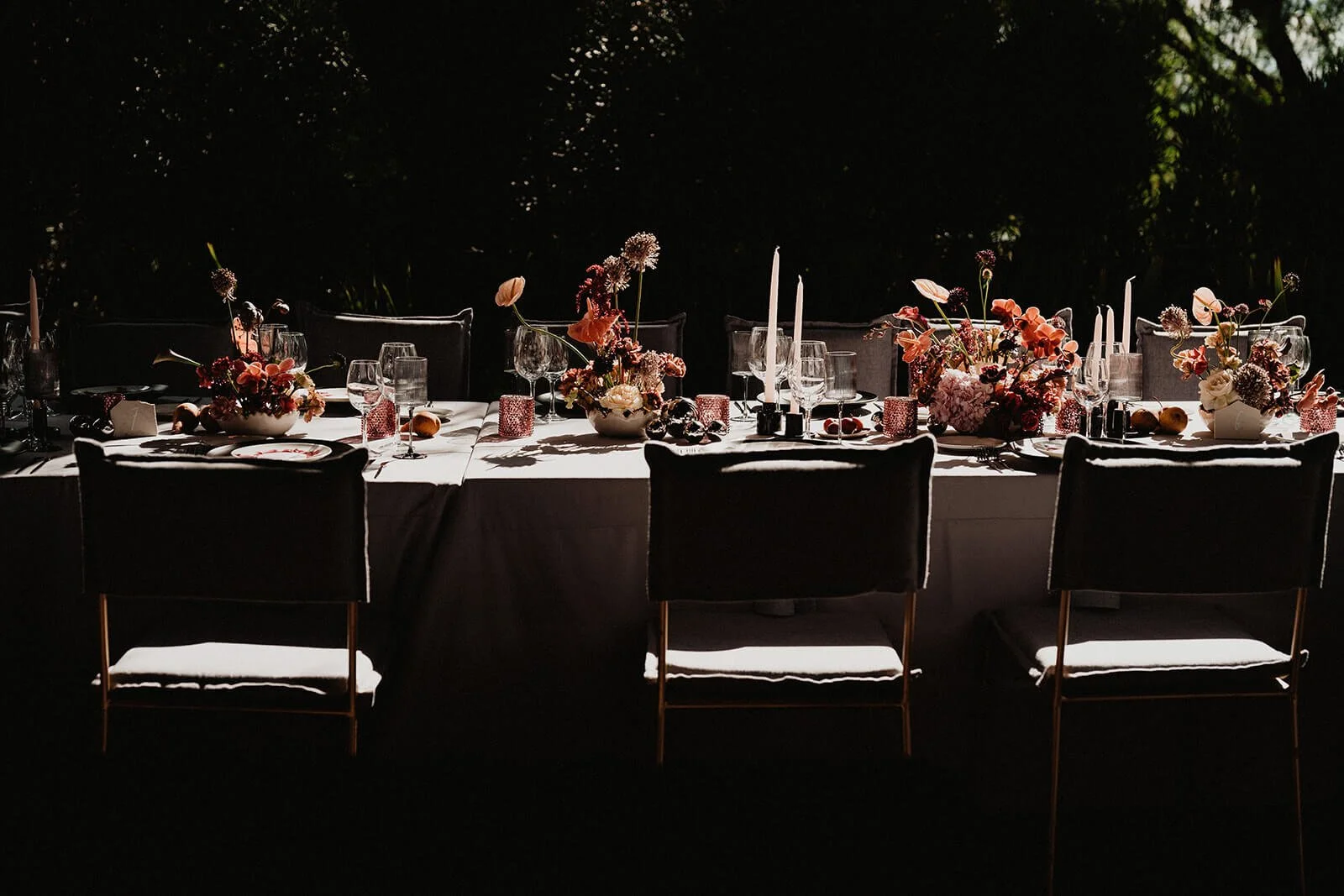 Elegant dining table set outdoors with flowers, candles, wine glasses, and place settings, illuminated by low lighting at night.