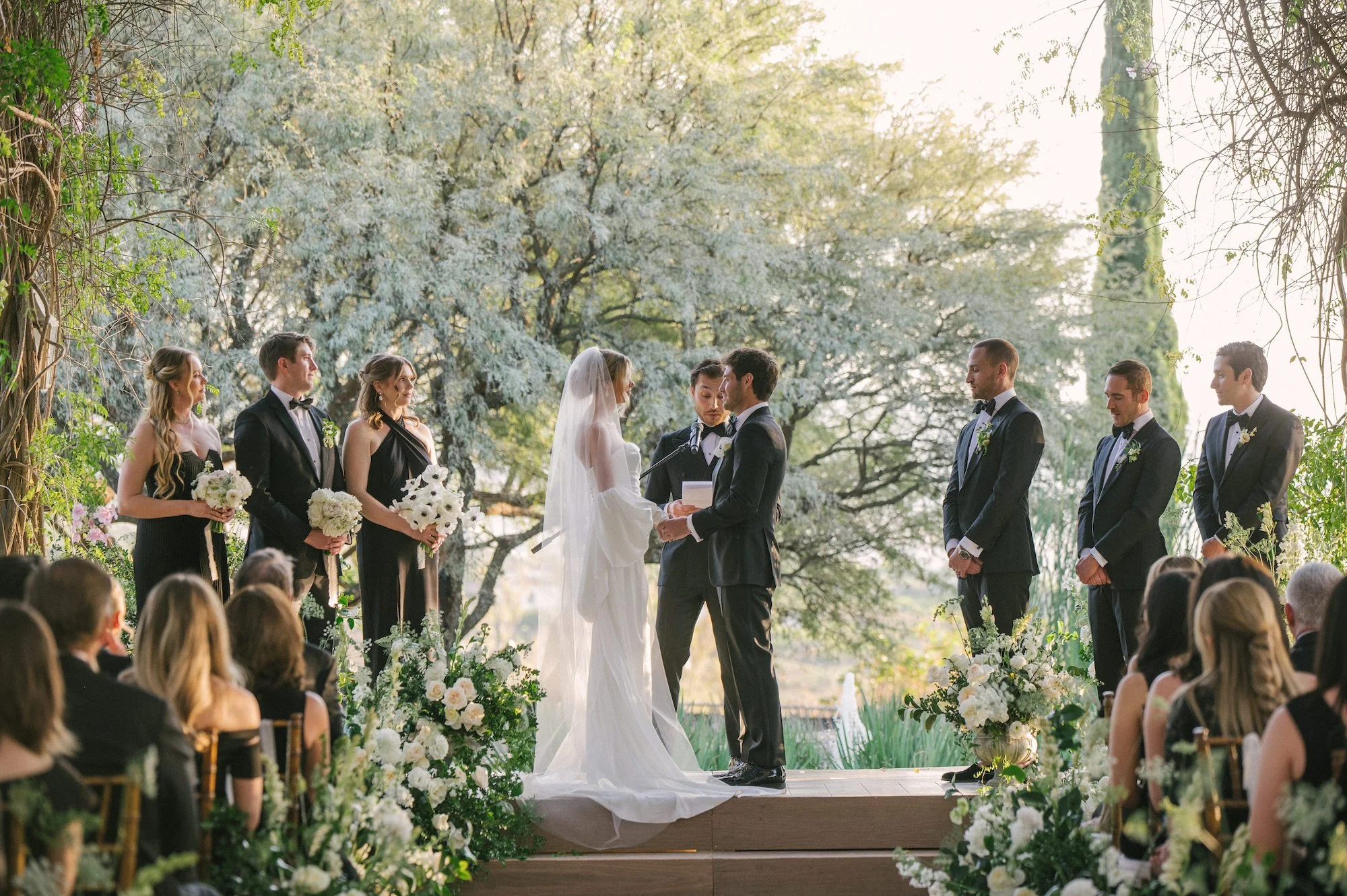 A wedding ceremony held outdoors with the bride and groom exchanging vows on a wooden platform surrounded by floral decorations and greenery, with guests seated nearby and bridesmaids and groomsmen standing on each side.