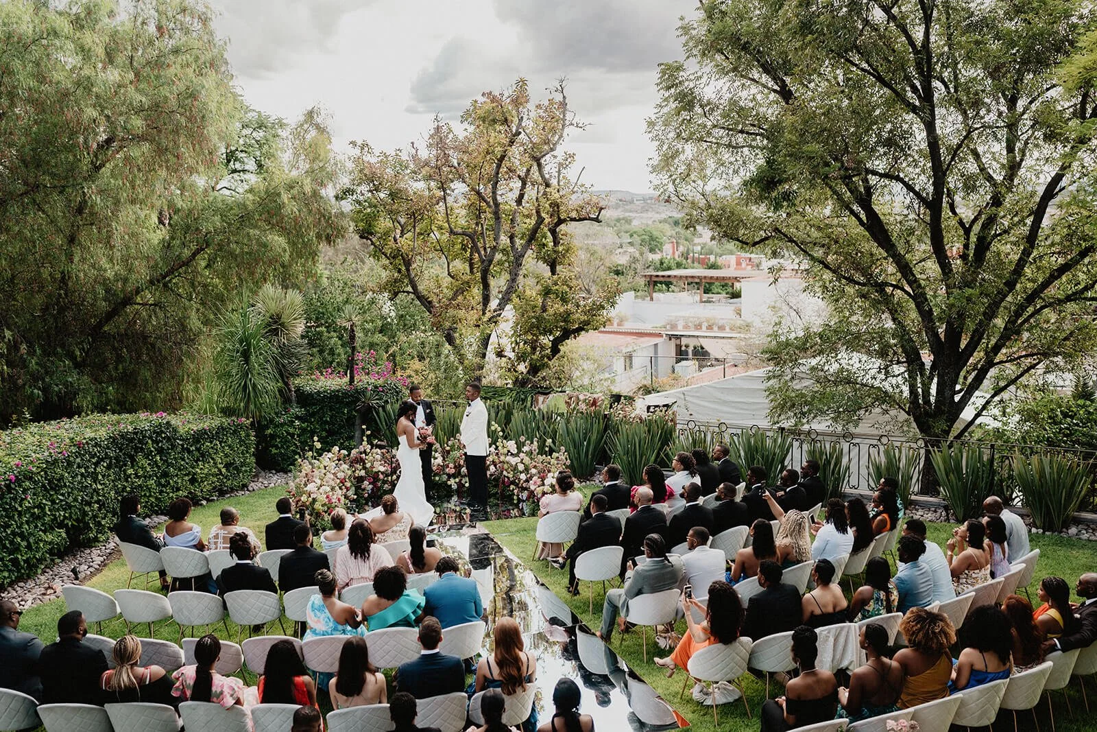 A wedding ceremony with a bride and groom standing at an altar surrounded by lush garden and guests seated on white chairs outdoors.