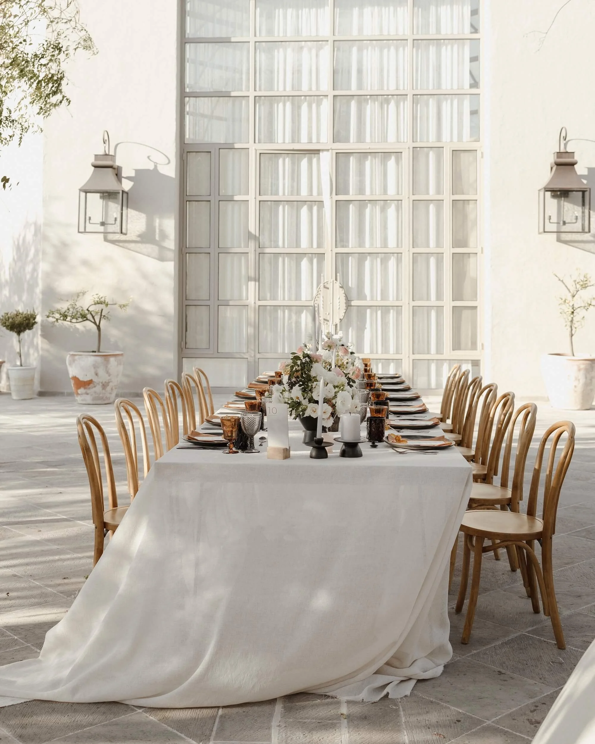 Elegant outdoor dining table set with white tablecloth, floral centerpiece, candles, and glassware, surrounded by wooden chairs and potted plants on a sunny patio with large window backdrop.