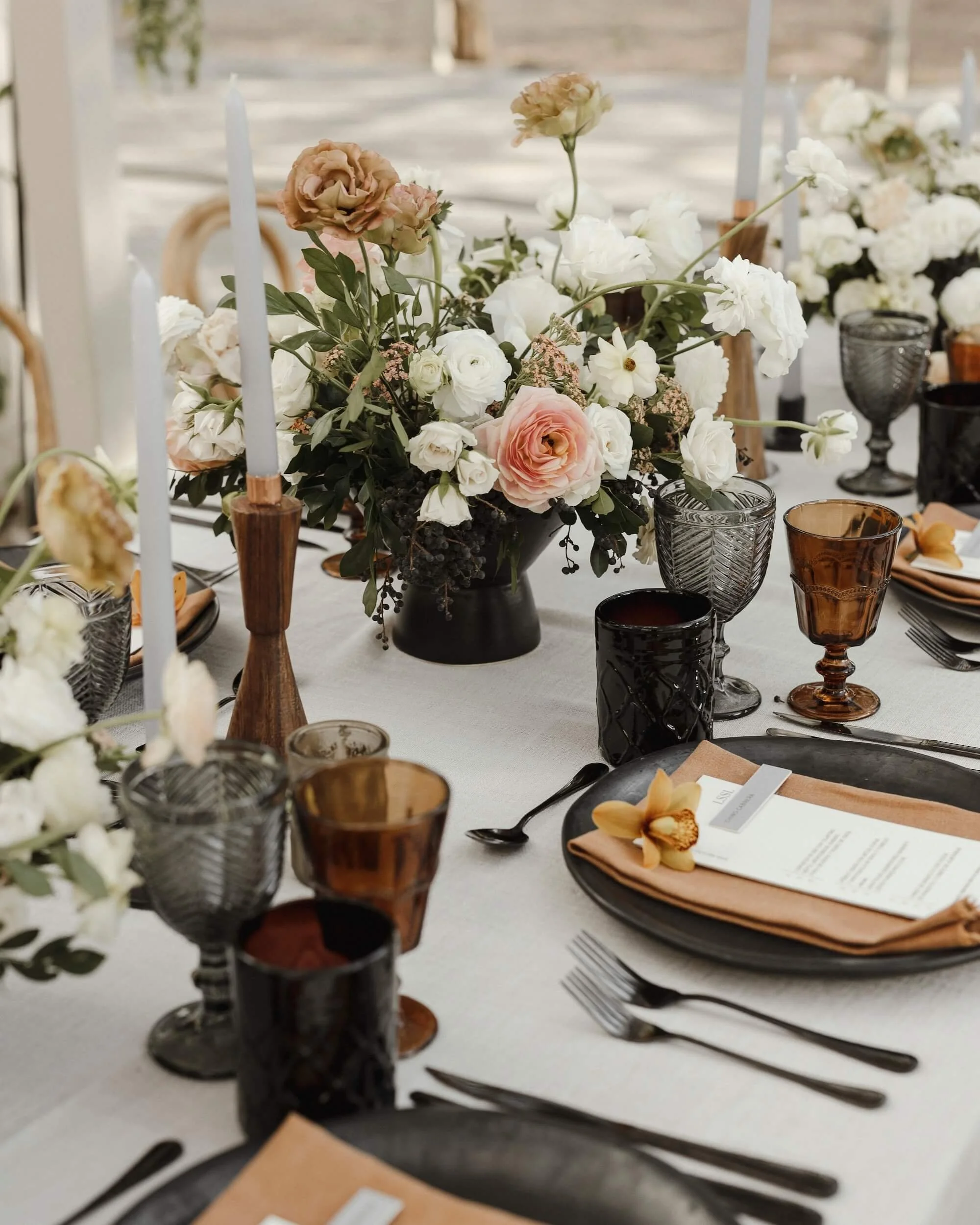 Elegant table setting with a floral centerpiece of pink and white flowers in a black vase, surrounded by amber and gray glassware, black plates, silverware, and peach-colored napkins with a day setting.