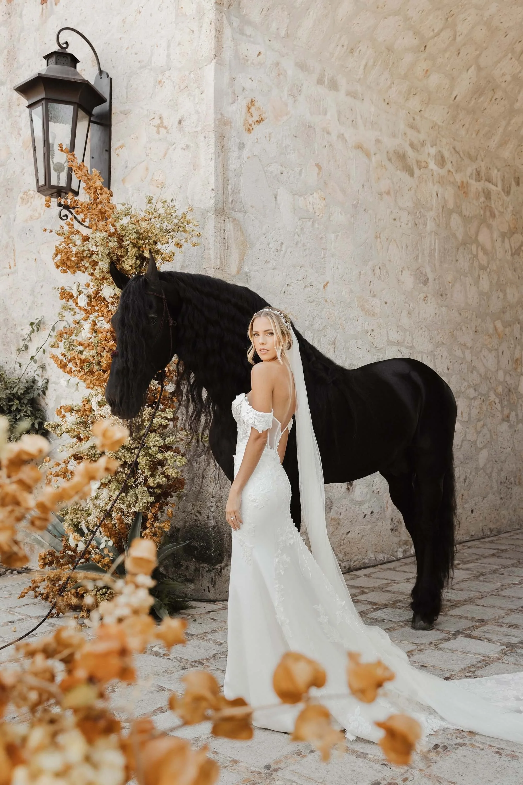 A bride in a white wedding gown with an open back and lace details, holding a long train, stands beside a black horse with a long, wavy mane, against a stone wall and decorated with flowers and a vintage lantern.