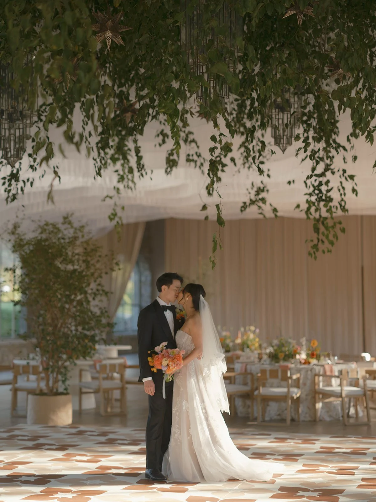 A bride and groom standing close together on the dance floor at their wedding reception, sharing a tender moment with their foreheads touching, in a decorated hall with large chandeliers, plants, and floral arrangements.