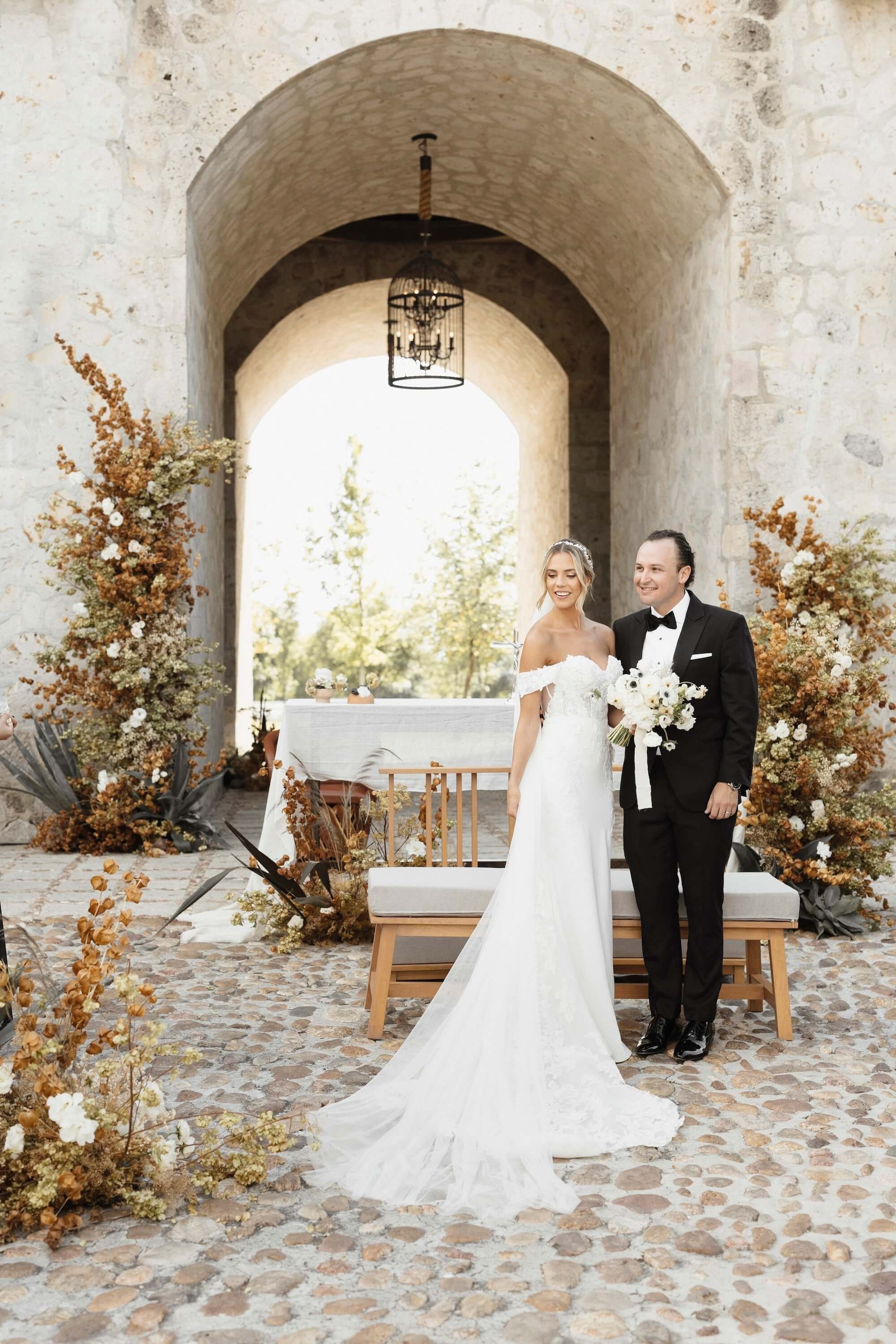 A bride and groom standing together under an arched stone structure with floral arrangements, smiling, during a wedding ceremony.