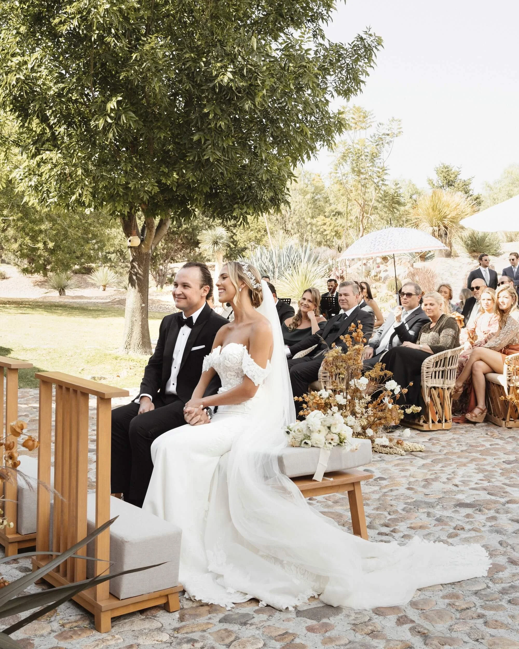 Bride and groom seated outdoors during their wedding ceremony, surrounded by seated guests on a sunny day with trees and plants in the background.