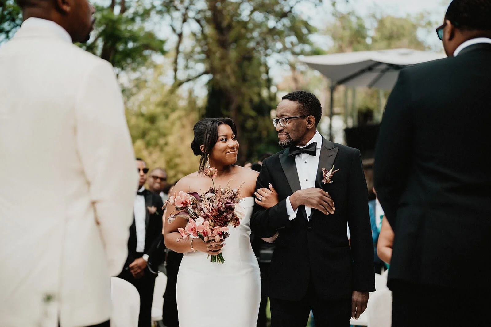 A bride in a white wedding dress holding a bouquet looking at a groom in a black tuxedo during an outdoor wedding ceremony.