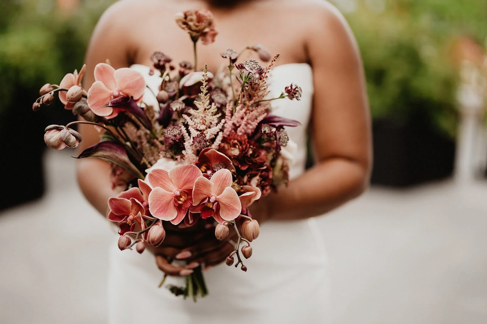 A woman in a white strapless dress holding a bouquet of pink and purple flowers outdoors.