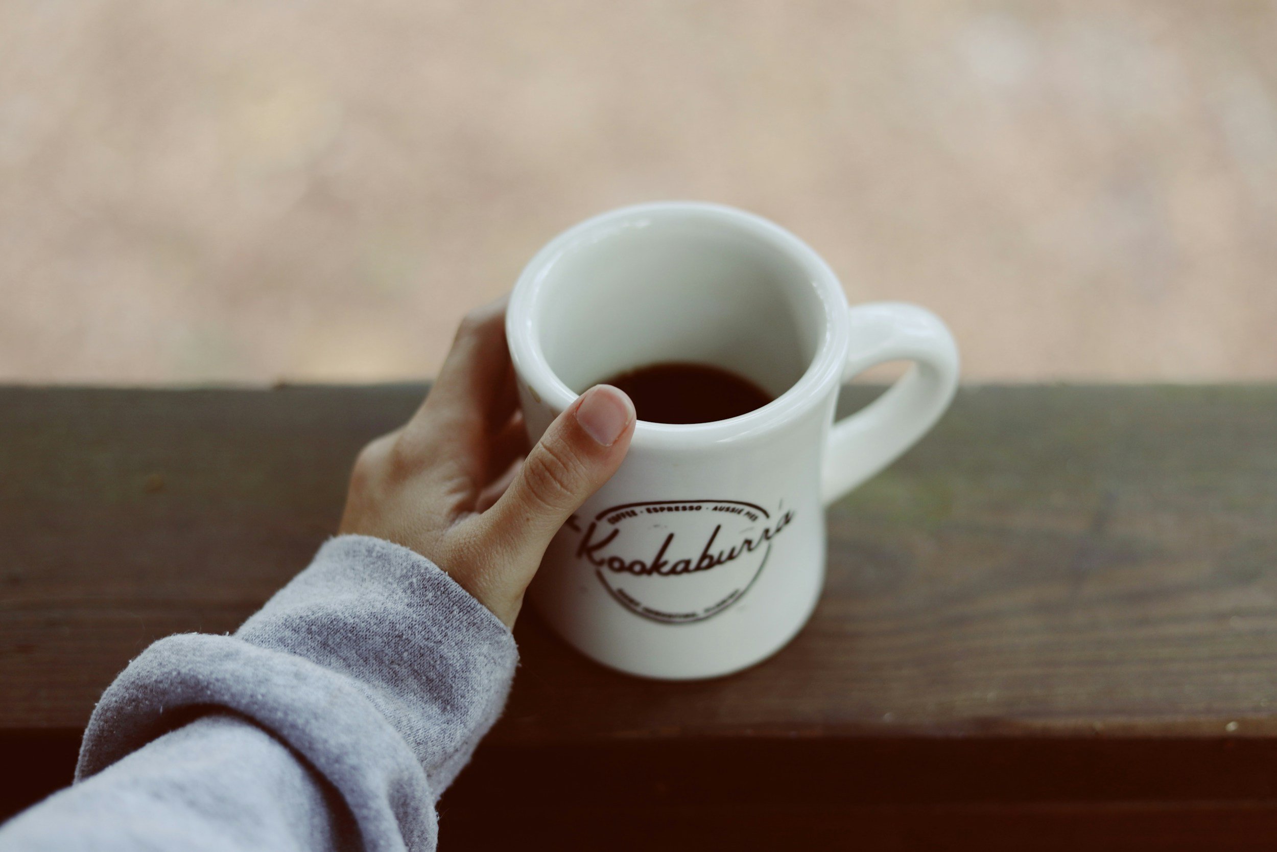 Person holding a white coffee mug with the word 'Kookaburra' printed on it, filled with coffee, sitting on a wooden surface.