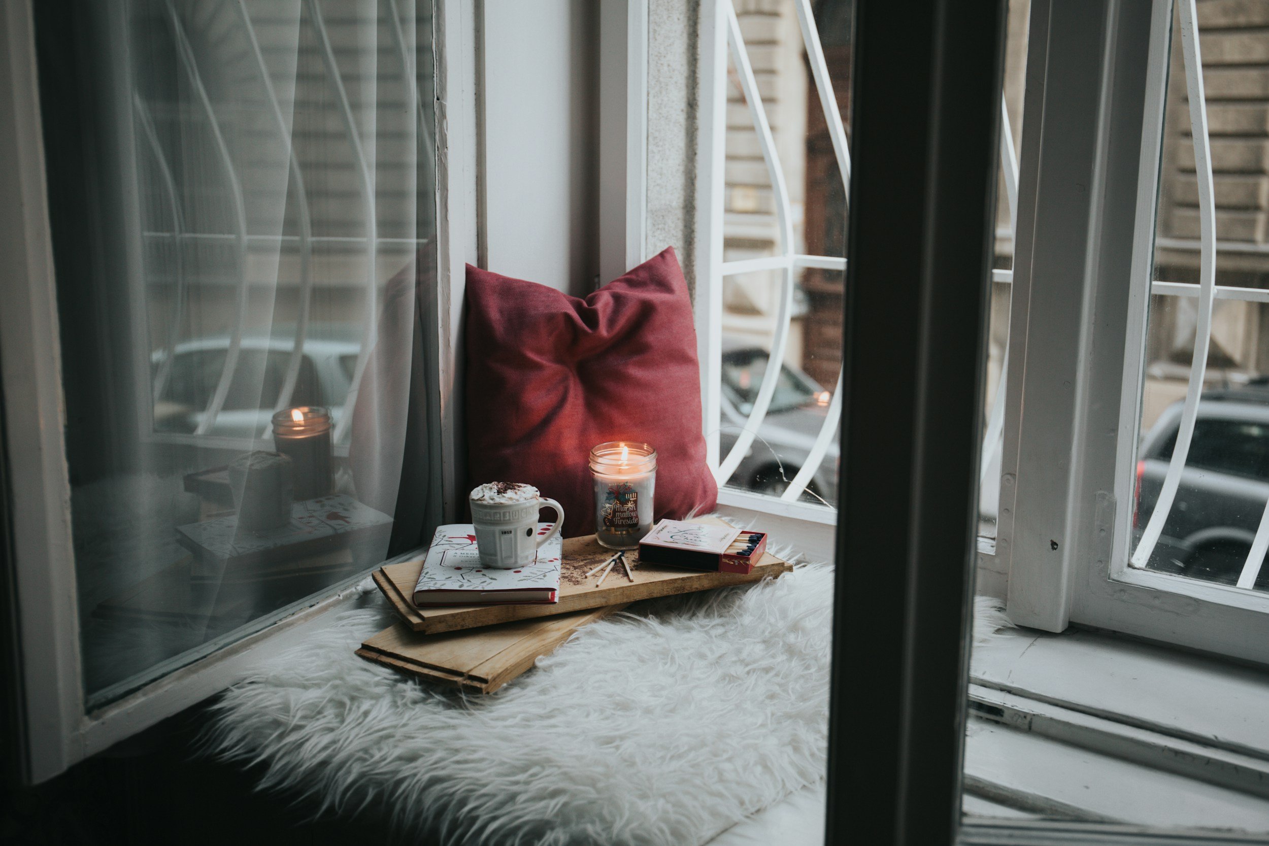 A cozy window nook with a fluffy white rug, a red pillow, a lit candle in a glass jar, a mug of hot chocolate with whipped cream, a small box of chocolates, and a few notebooks placed on a wooden tray, with a view of parked cars outside.