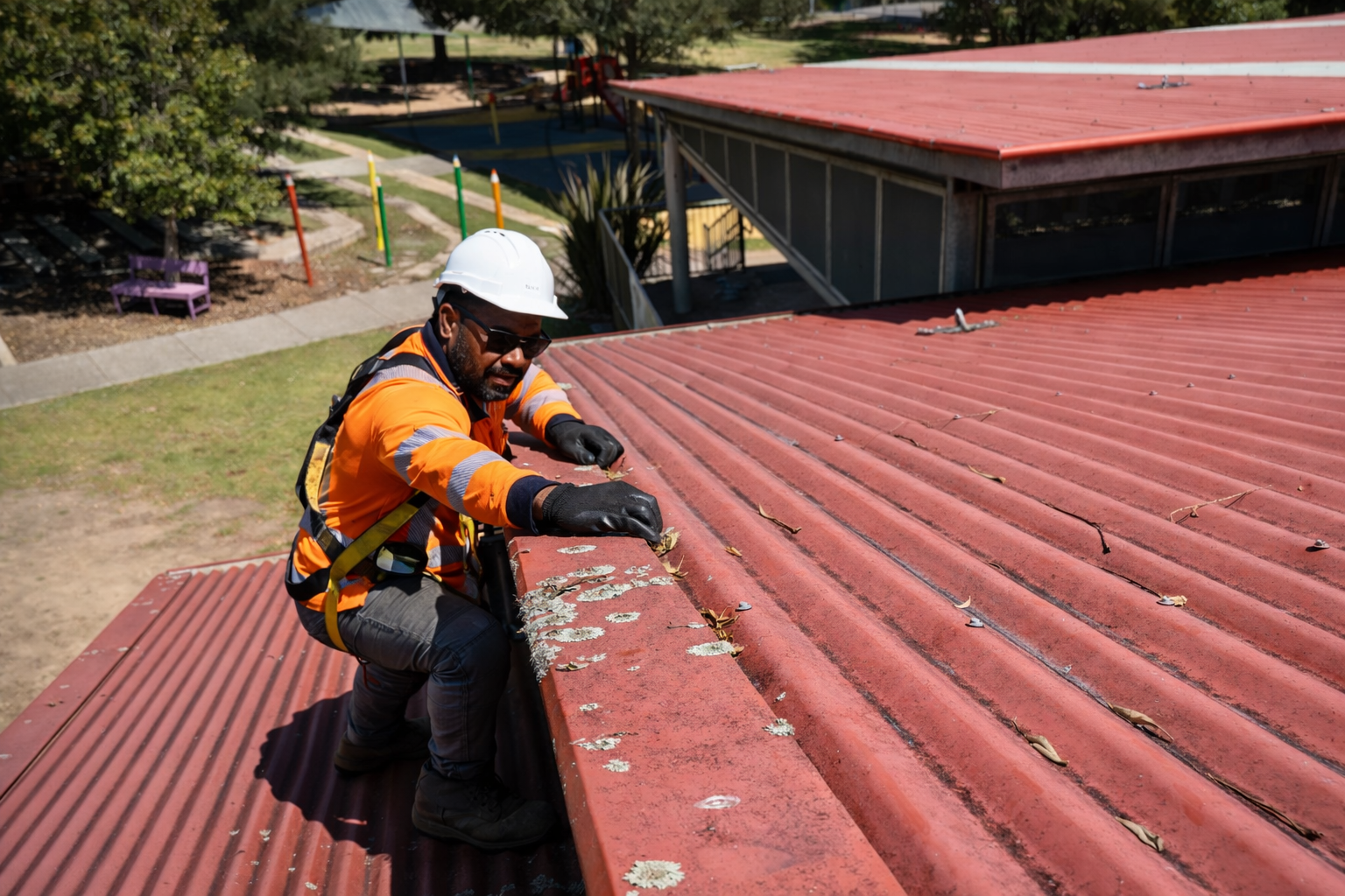 A worker wearing a white safety helmet, sunglasses, orange safety jacket, gloves, and a safety harness kneels on a red metal roof, inspecting or repairing it with a tool, while looking down at the roof surface.