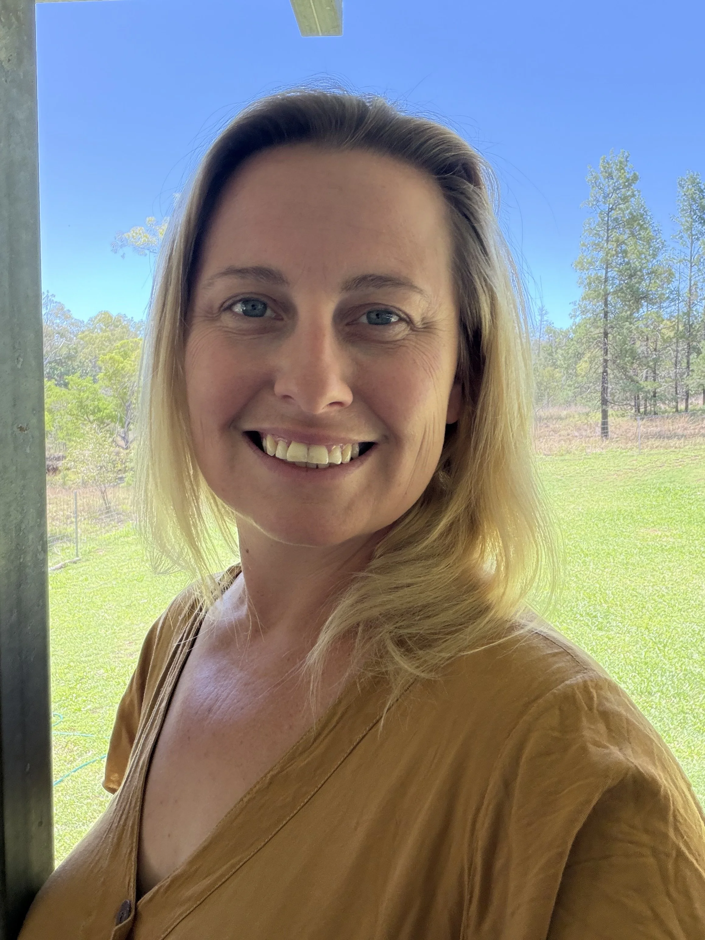 A smiling woman with blonde hair and blue eyes standing outdoors near a window, with trees and a blue sky in the background.