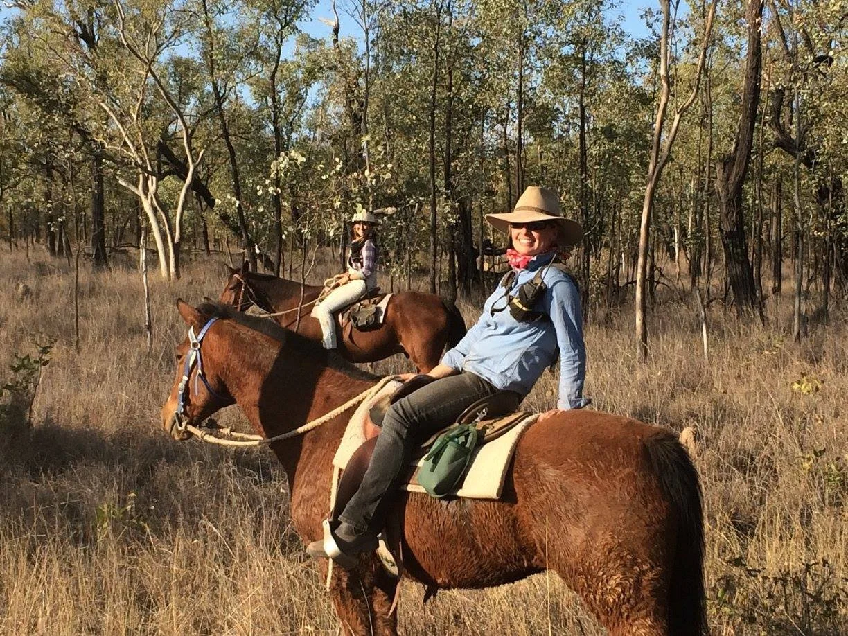 Two women horseback riding in a forested area with dry grass and trees, bright sunlight, wearing hats, shirts, and riding gear.