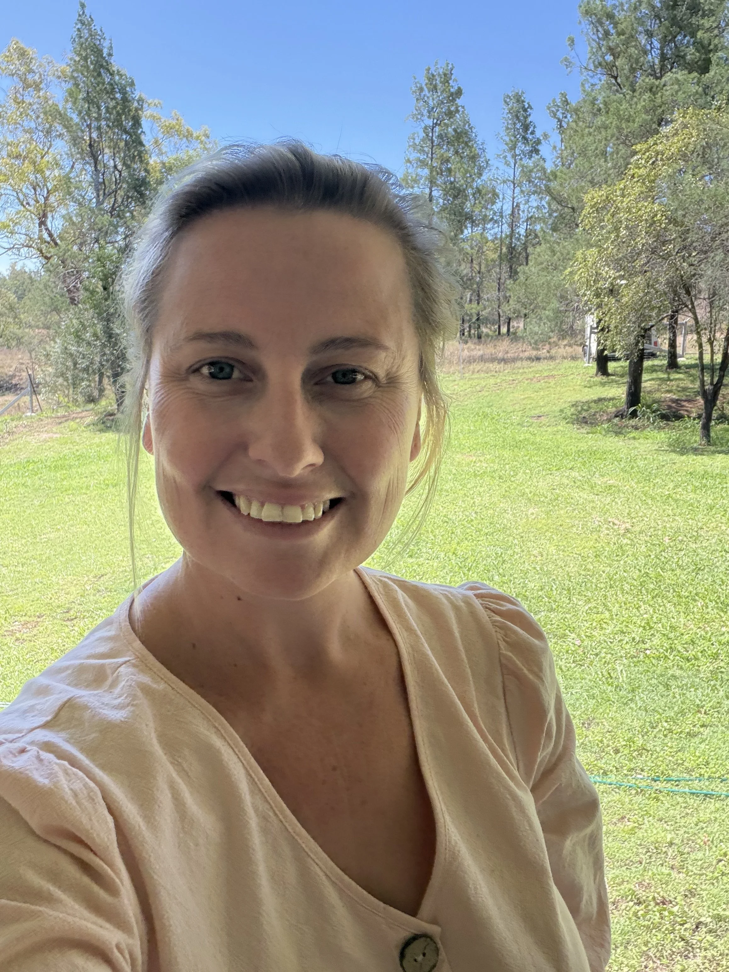 A woman smiling outdoors on a bright, sunny day with a background of green grass, trees, and a blue sky.