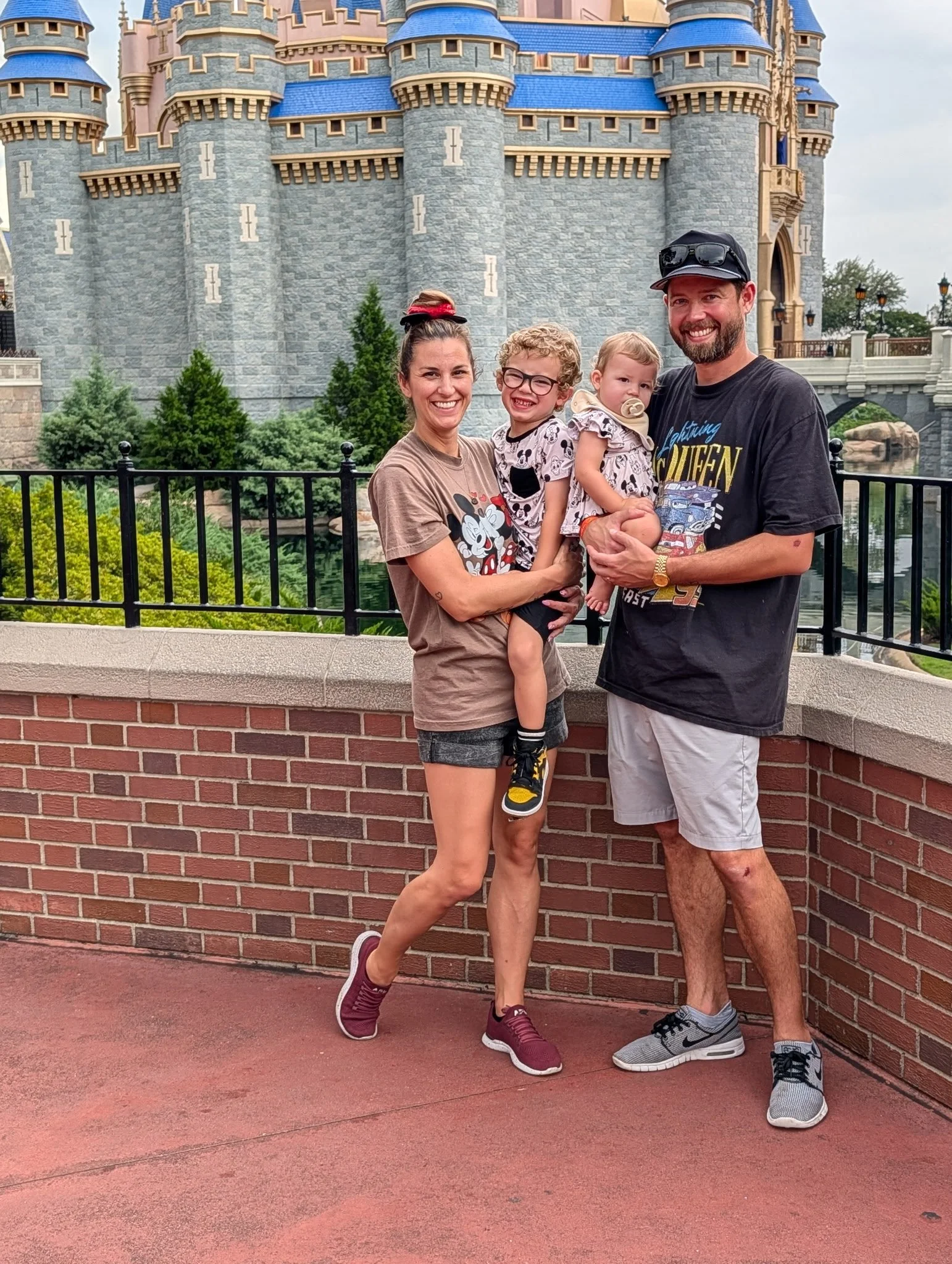 A family of four standing in front of a castle at Disneyland, with the mother holding a young girl, and the father holding a toddler. The castle is gray with blue rooftops, and there is greenery and a black fence behind them.