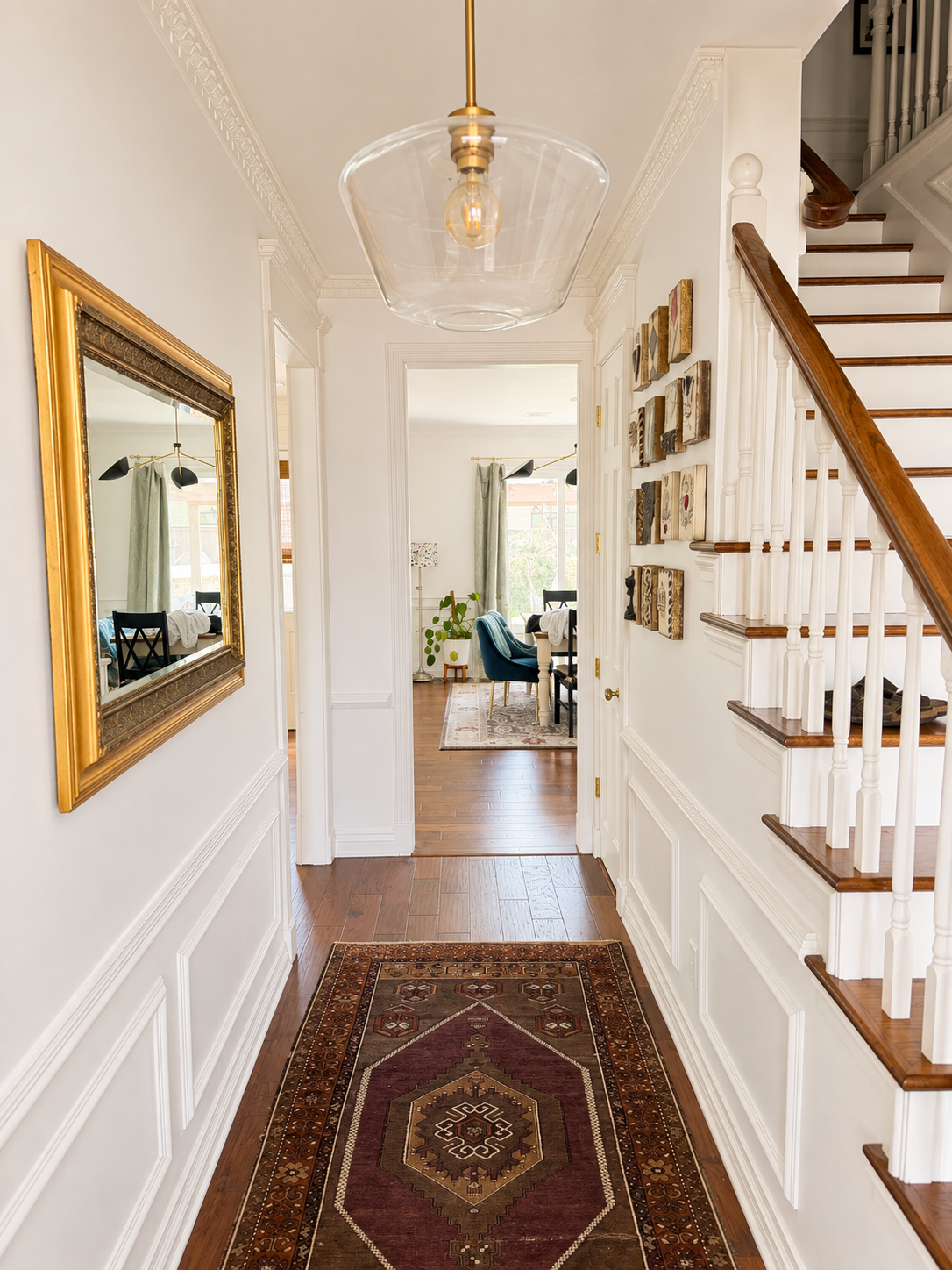 Hallway with a wooden floor, white walls, and a patterned rug, overlooking a dining room with windows, curtains, chairs, and a small table, and a staircase with wooden treads and white spindles on the right.