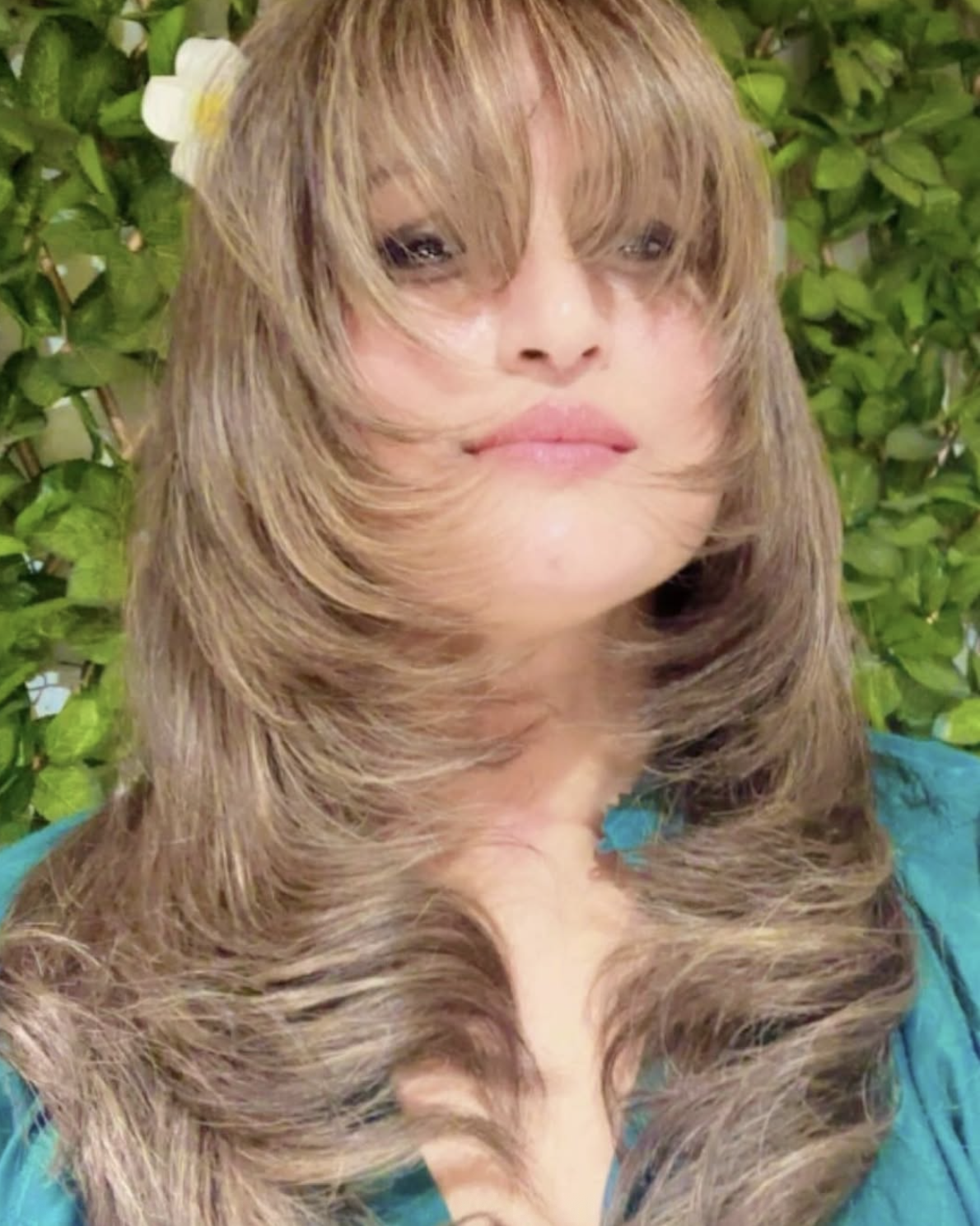 A woman with long, wavy light-brown hair partially covering her face, standing in front of green foliage with a white flower in her hair.