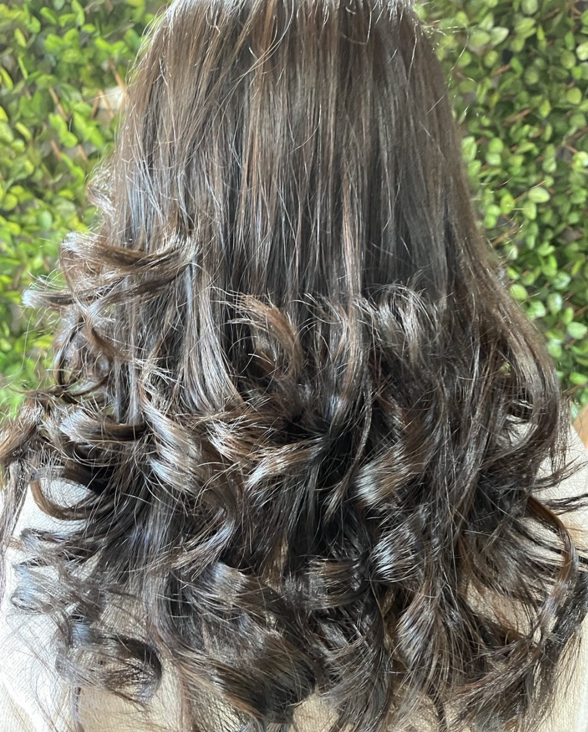 Back view of a person with long, dark brown, wavy, and curly hair, standing in front of a green leafy background.