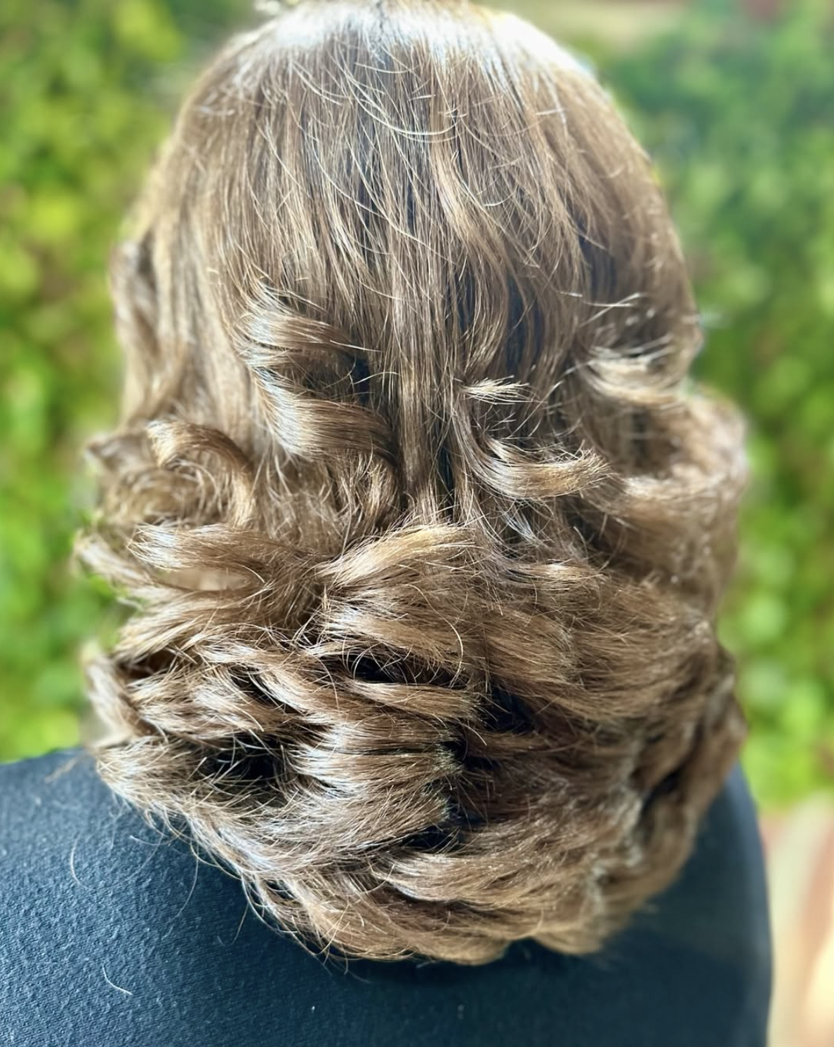 Back of a person's head with curly, light brown hair outdoors with green foliage in the background.