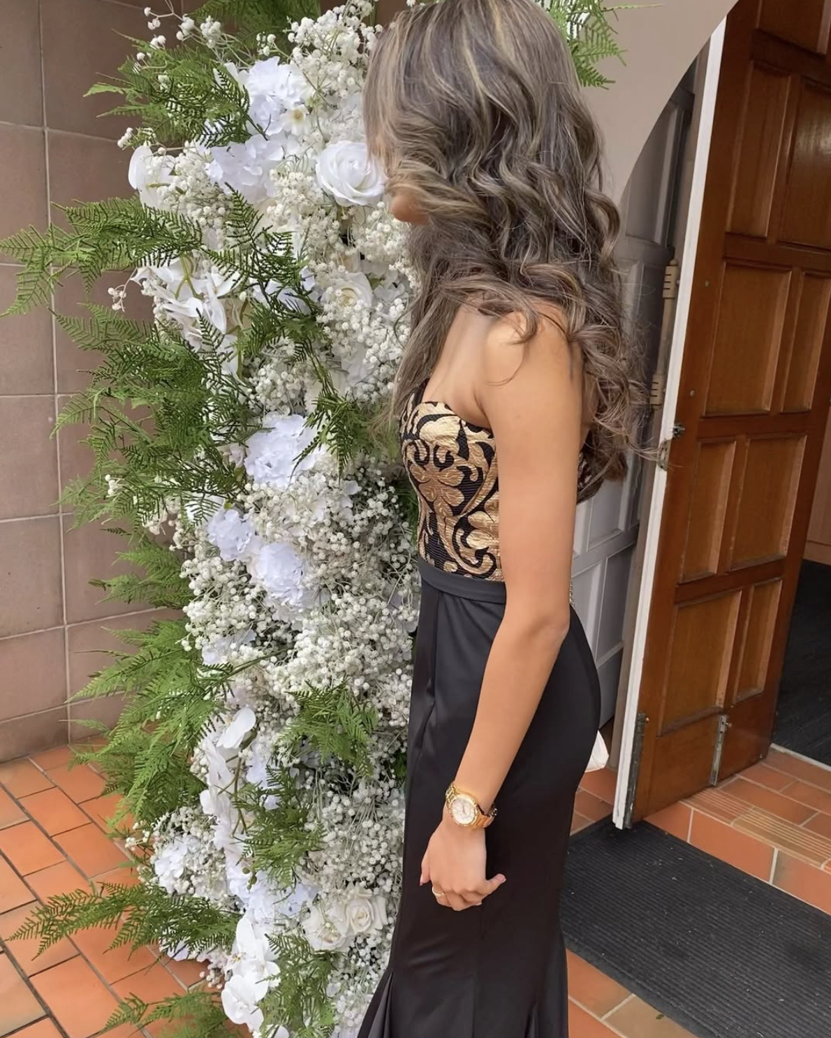 A woman with long, curly, brown hair in a strapless black and gold dress stands beside a large floral arrangement of white flowers and green foliage in an indoor setting.