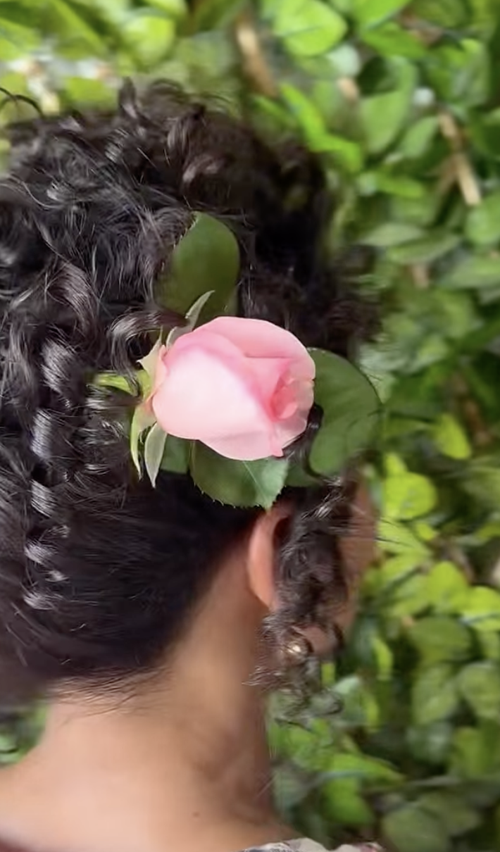 A woman with curly black hair has a pink flower tucked behind her ear while standing among green leaves.