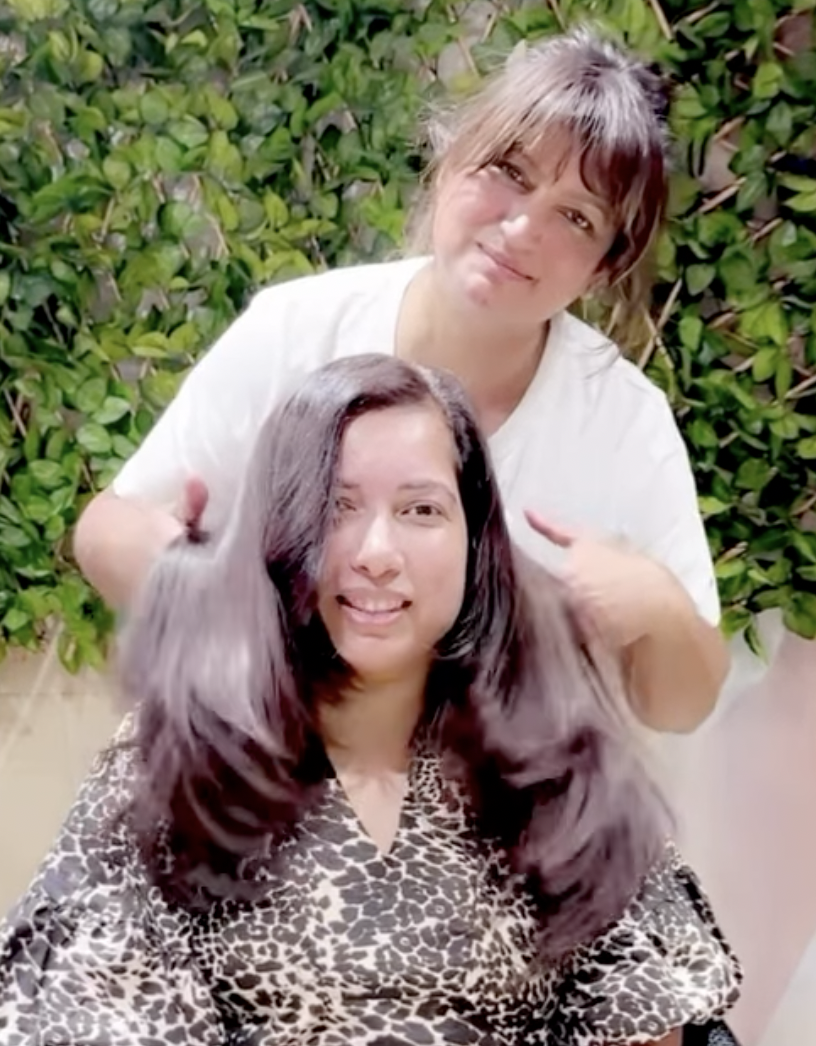 Two women, one standing behind the other, smiling. The woman in front has long dark hair and is wearing a leopard-print top. The woman behind has shoulder-length light brown hair and is wearing a white shirt. They are outdoors with green leafy bushes in the background.